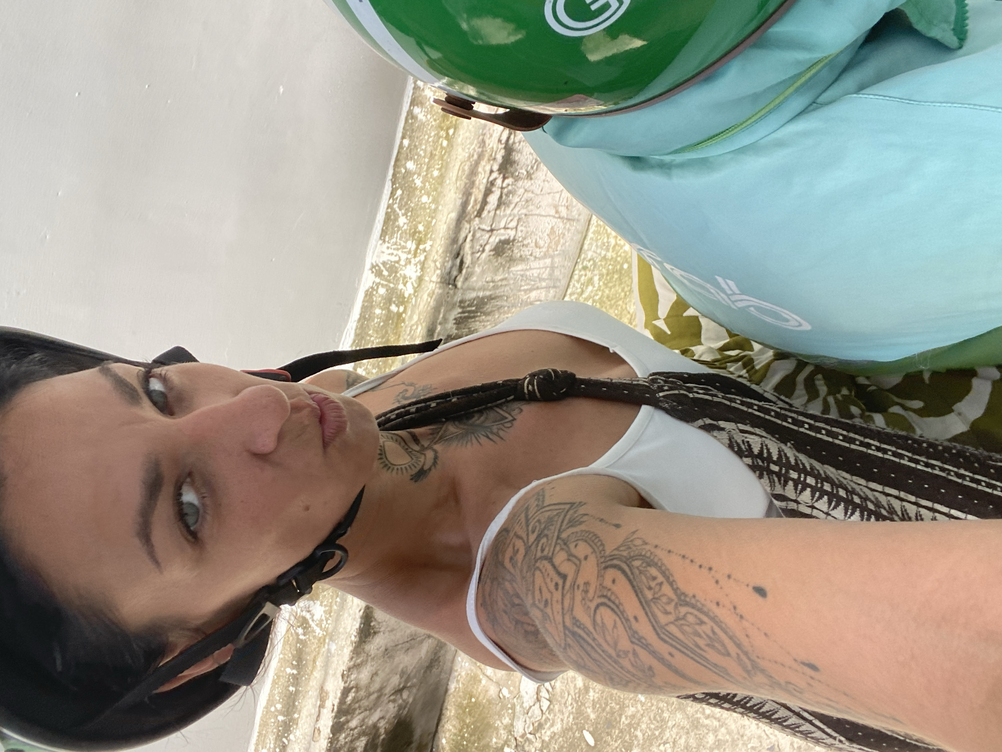 The image is a selfie taken by a woman wearing a black helmet and a white tank top. She has extensive tattoos on her left arm and chest, and a black strap from her bag crosses her chest. She is looking directly at the camera with her lips pursed. To her right, another person is wearing a light blue jacket and a green helmet. The background appears to be an outdoor setting with stone steps leading to a body of water. The overall impression is of someone about to embark on or having just completed a scooter or motorcycle ride. While the exact location in Phu Thuy Ward, Vietnam, is not explicitly identifiable from the image, the presence of scooters, helmets, and the general aesthetic suggests a Southeast Asian context.