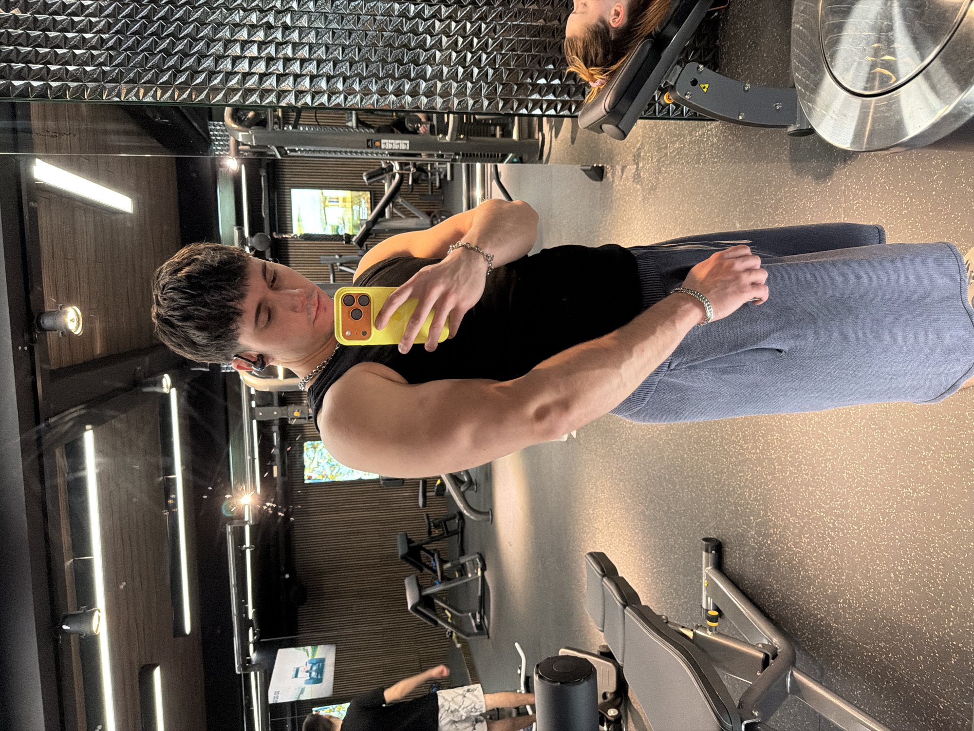 A man with dark hair is taking a selfie in what appears to be a gym. He is wearing a black tank top and blue sweatpants and has a yellow phone in his hand. His reflection is visible in a mirror, showing the gym environment with various exercise machines. In the reflection, other individuals can be seen in the background. Based on the gym's appearance and available data, it may be located in Sofia, Bulgaria, but this is not definitively verifiable from the image alone.