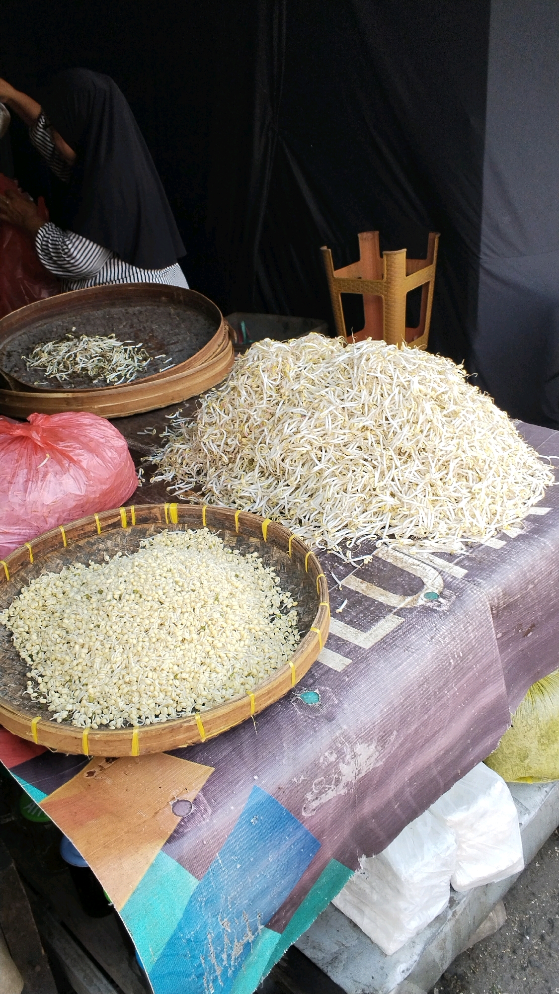 In Mojokerto, Indonesia, a market scene unfolds under a black tarp. A person wearing a black hijab and a striped shirt is partially visible, tending to a large pile of bean sprouts. In front of the bean sprouts, a woven basket contains a heap of smaller, lighter-colored sprouts. To the left, a red plastic bag sits next to another woven tray, this one holding a smaller quantity of the same light-colored sprouts. Behind the bean sprouts, a wooden rack is visible. The scene takes place on a table covered with a patterned cloth, with stacks of white plastic-wrapped items in the foreground.
