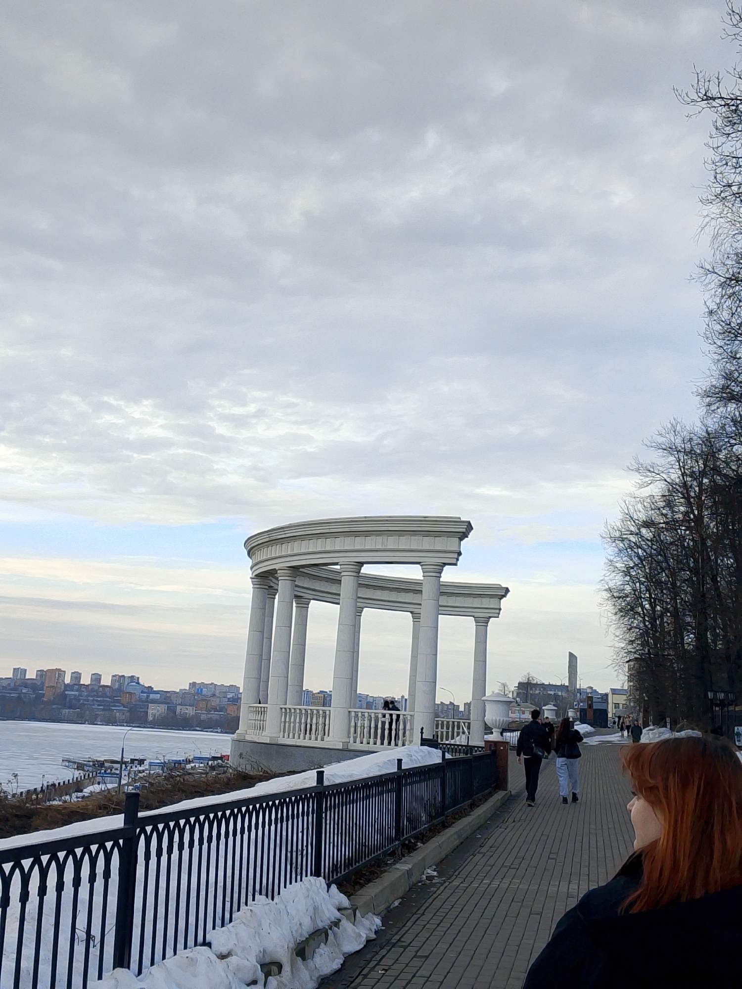 The image depicts a view along a pedestrian walkway in Izhevsk, Russia, on what appears to be a cold day. Patches of snow are visible along the base of a black wrought-iron fence that borders the walkway.
In the foreground, the back of a person with reddish-brown hair is visible on the right. Two people are walking along the path in the middle distance. Behind them, an open-air rotunda-like structure with white columns stands overlooking what appears to be a partially frozen body of water. In the background, across the water, are several buildings. The sky is overcast.