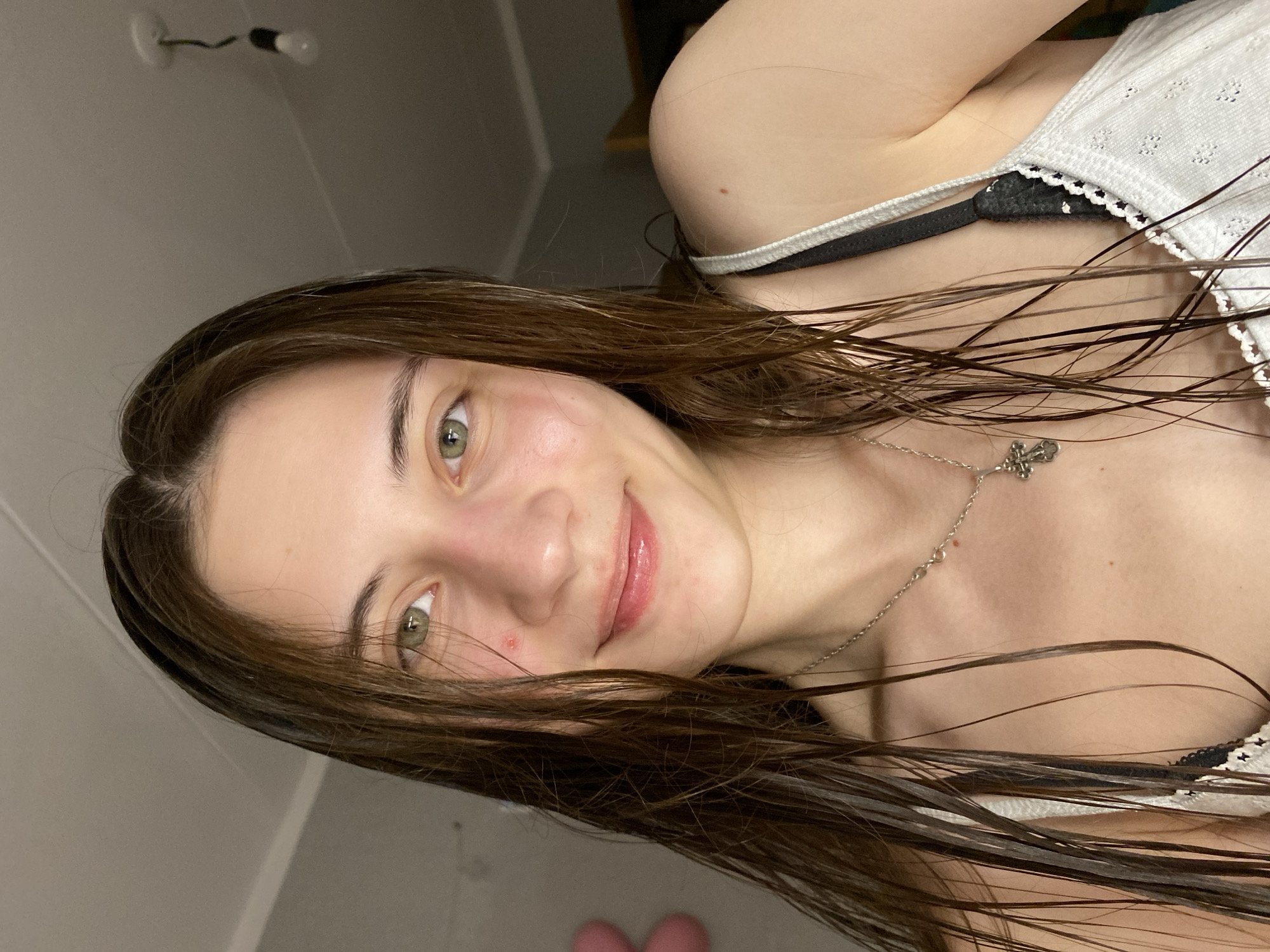 A close-up, selfie-style photograph depicts a young woman with wet, dark brown hair and light skin. She has light green eyes and a small red mark near her nose. She is smiling slightly. She is wearing a white top with a black bra visible underneath. A silver necklace with a cross pendant rests on her chest. The background is blurred, showing a light-colored wall and ceiling. In the top left corner, a light fixture is visible. The image does not contain sufficient information to determine a specific location in Votkinsk, Russia. There is no discernible evidence to suggest a specific location.
