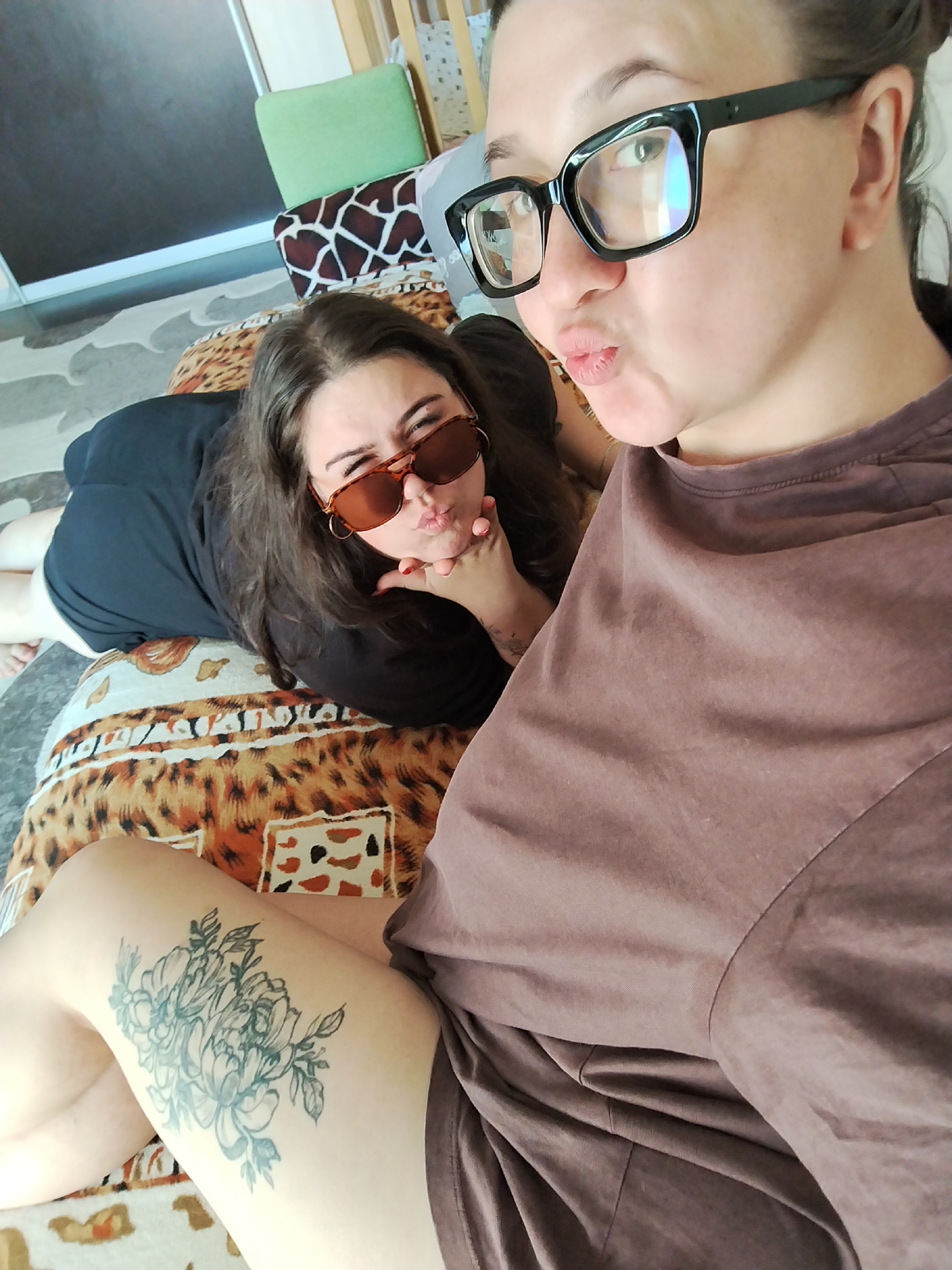 This is a medium shot, taken from above, of two women indoors. The woman in the foreground, on the right, is wearing black-rimmed glasses and a brown t-shirt. She is making a kissing face towards the camera. The woman in the background, on the left, is lying on her side and is also making a kissing face. She is wearing sunglasses and a black t-shirt. She has long, dark hair. Both women appear to be young adults. They are lying on a patterned surface, possibly a bed or sofa, which has a leopard print design. In the background, there is a crib with a green pillow and a giraffe-patterned cushion. The flooring visible is a light-colored carpet with a wavy pattern. There is also a tattoo of flowers on the left thigh of the woman in the foreground. The image does not contain any specific clues about its location in Chaikovsky, Russia.