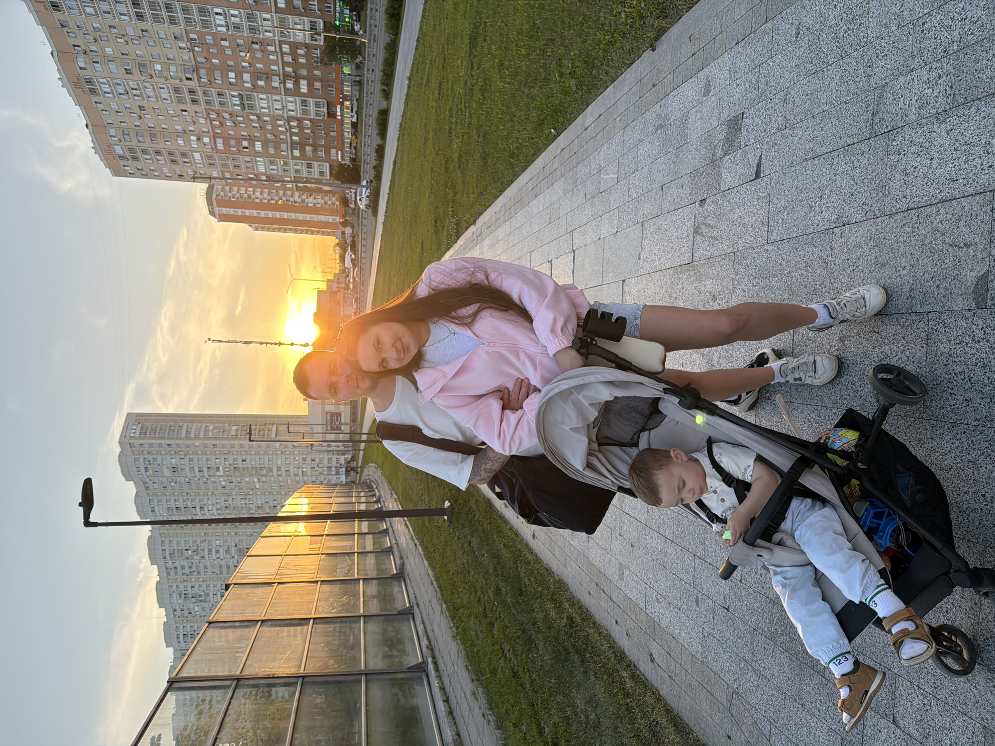 A family of three, consisting of an adult male, an adult female, and a young child in a stroller, stands on a grey paved pedestrian path in an urban environment. The scene is illuminated by the warm, golden light of a low sun, likely at sunset, visible on the horizon between tall buildings.
The man, positioned slightly behind the woman on the left, wears a white short-sleeved t-shirt, dark pants (partially obscured), and carries a black duffel bag over his left shoulder. Tattoos are visible on his right arm. He has short dark hair and light stubble, looking towards the camera. The woman, standing slightly in front of him, wears a pink zip-up hoodie over a light gray t-shirt, light denim shorts, and white sneakers. She has long brown hair parted in the middle and smiles subtly at the camera, with her left arm around the man's waist and her right hand resting on the handle of the stroller.
The young child, a boy with light brown hair, sits in a grey stroller, secured by a harness. He wears a white patterned short-sleeved shirt, light blue pants, and brown sandals over white and green striped socks. His gaze is directed downwards at a small, light-colored object held in his hands. The stroller features a black frame, a dark storage basket underneath containing blue and yellow items (likely toys), and a black cup holder attached to the right handle.
To the left of the path, a transparent or translucent multi-paneled sound barrier reflects the bright orange sunlight intensely. Behind this barrier, several tall, light-colored, multi-story apartment buildings with numerous windows are visible. A dark streetlamp stands between the barrier and the path. To the right of the path, a wider strip of mown grass separates the walkway from a multi-lane asphalt road with cars. Beyond the road, more tall residential buildings, some with balconies, line the street. Visible on the ground level of these buildings are commercial signs in Cyrillic, including "ПЯТЕРОЧКА" (Pyaterochka, a supermarket chain) and "ГОРЗДРАВ" (Gorzdrav, a pharmacy chain), indicating a location in Russia, specifically Moscow given the architectural style and common retail presence. The sky above is mostly clear with some wispy clouds, showcasing the vibrant sunset colors.