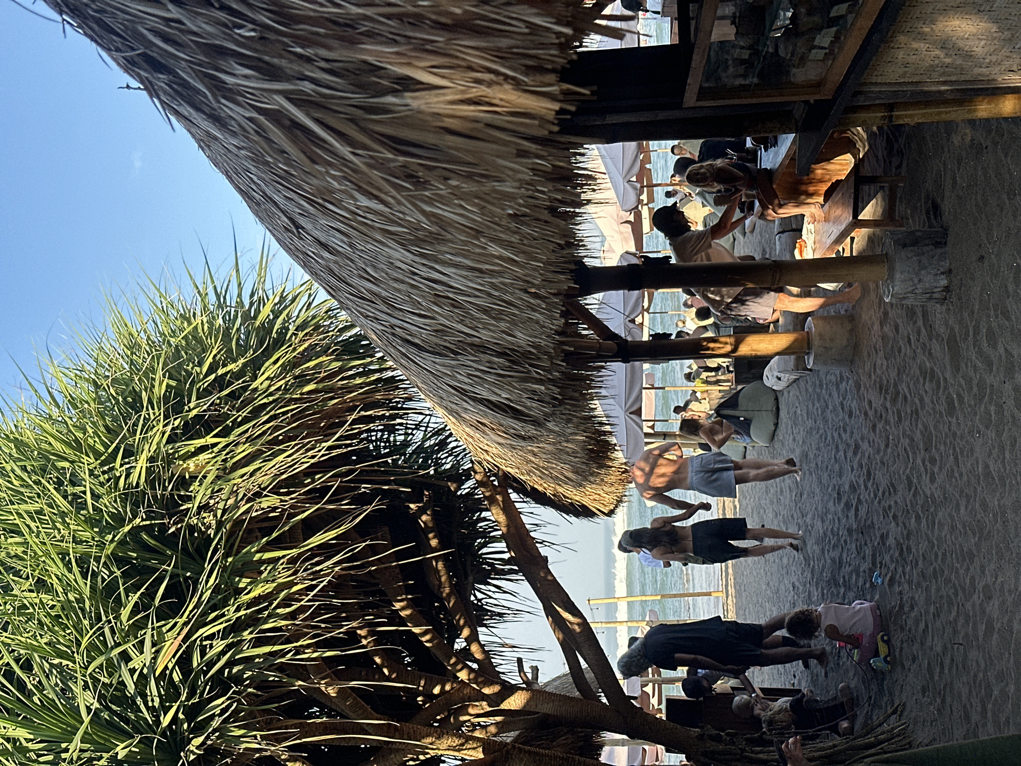 This image captures a vibrant beach scene in North Kuta, Indonesia, likely during the late afternoon. The foreground features dark sand, with a large, leafy, pandanus-like tree dominating the left side, casting significant shadows. Its complex root system and branches are visible. To the right, a large thatched roof structure, supported by wooden posts and concrete bases, extends into the mid-ground, providing shade.
Numerous individuals populate the scene. In the mid-ground, a child in a pink top sits on the sand, playing with a pink and yellow toy truck and a small blue object. Near the tree, a man with long grey hair in a black t-shirt stands facing right. A couple walks hand-in-hand towards the ocean; the man is shirtless in grey shorts, and the woman wears a dark bikini top and dark shorts. To their right, a grey beanbag chair rests on the sand. Further right, beneath the thatched roof, a man in a white t-shirt stands near a young blonde girl sitting on a wooden bench, appearing to hand her something. A display case with multiple shelves is partially visible on the far right.
In the background, the ocean features moderate waves under a clear blue sky. White or light-colored beach umbrellas are scattered along the shoreline, shading tables and numerous other people enjoying the beach environment. The overall atmosphere suggests a casual beach club or restaurant.