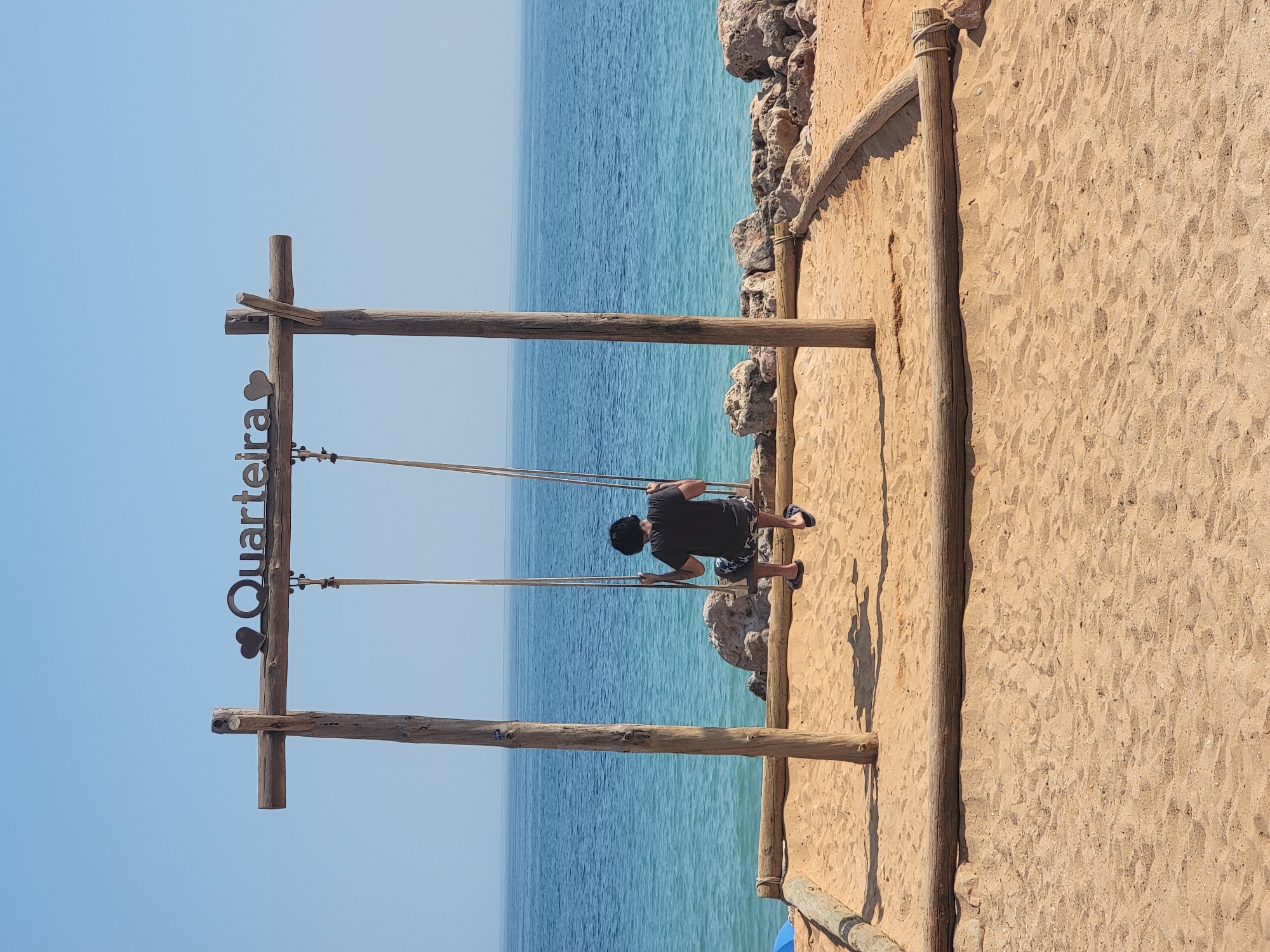 The image shows a person on a swing overlooking the ocean. The swing set is made of rustic wooden posts and beams, with ropes holding the swing seat. A metal sign on the left post reads "Quarteira" with a heart symbol. The person, wearing a black t-shirt and patterned shorts, is seated on the swing, facing away from the camera and towards the sea.
The scene is a sandy beach with the blue ocean stretching out to the horizon. Rocky formations are visible along the edge of the beach. The sky is clear and bright blue, indicating a sunny day, likely midday given the strong shadows. The overall mood is relaxed and picturesque, suggesting a vacation or leisure activity at a coastal location. The sand is textured with footprints and the natural undulations of the beach. The wooden structure is weathered, adding to the natural, beach-like aesthetic.