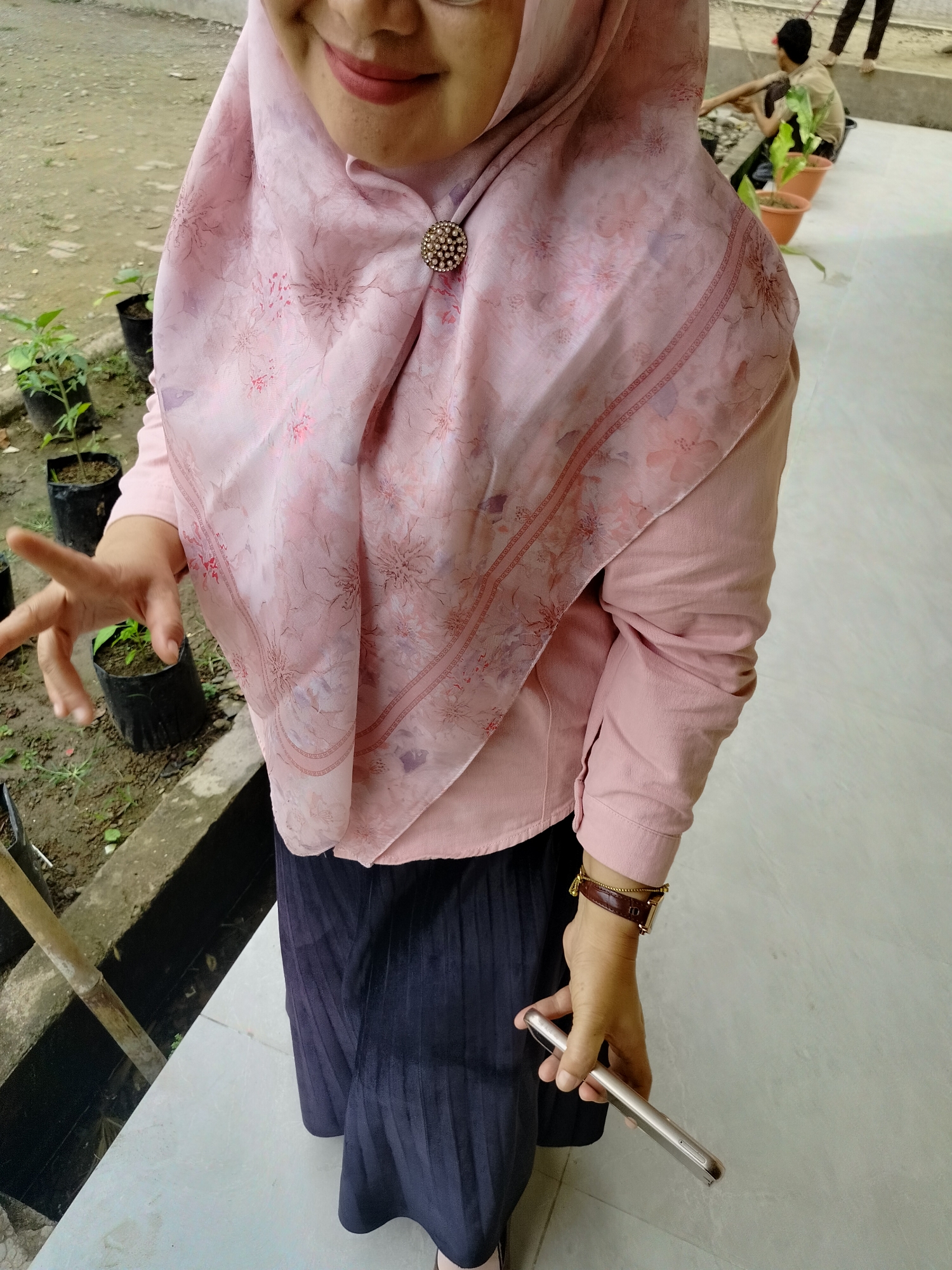 In Lawebekung, Indonesia, a woman wearing a pink hijab with a floral pattern and a delicate brooch stands on a tiled walkway. She is dressed in a pale pink long-sleeved shirt and a dark, pleated skirt. Her right hand, adorned with a brown leather watch, holds a silver smartphone. In the background, several small plants in black plastic bags are visible, along with another person tending to potted plants. The ground appears to be dirt and gravel.