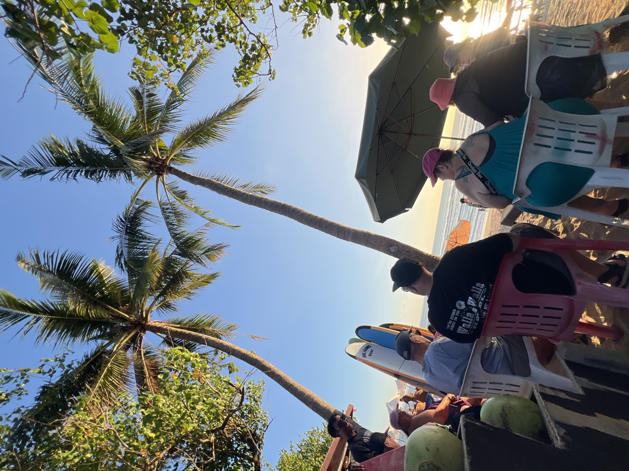 A group of people are seated on chairs on a sandy beach in Kuta, Indonesia. Two tall palm trees are visible in the foreground, with their fronds extending towards a clear blue sky. Several surfboards are propped up behind some of the seated individuals. A large green umbrella provides shade for some of the people. In the background, the ocean stretches out to the horizon, with gentle waves breaking on the shore. The sun appears to be setting, casting a warm glow over the scene. One person is standing near a wooden structure, seemingly looking out at the ocean. In the foreground, two green coconuts are placed on a dark wooden surface. The overall atmosphere is relaxed and tropical.
