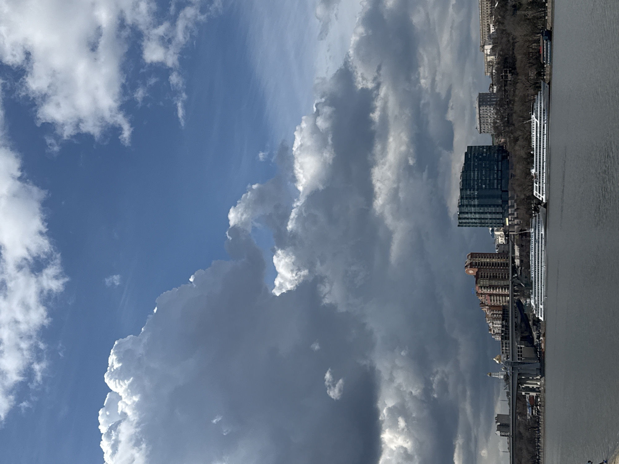 The image shows a view of Rostov-on-Don, Russia, with a focus on the cityscape and the sky. The buildings are dense and varied in architectural style, including modern glass structures and older, more traditional designs. A river runs along the edge of the city, with a large cruise ship docked along the embankment. In the sky, there are formations of clouds, ranging from bright white to dark grey, indicating potentially stormy weather. The sky contrasts with patches of clear blue. There are no people visible in the shot.