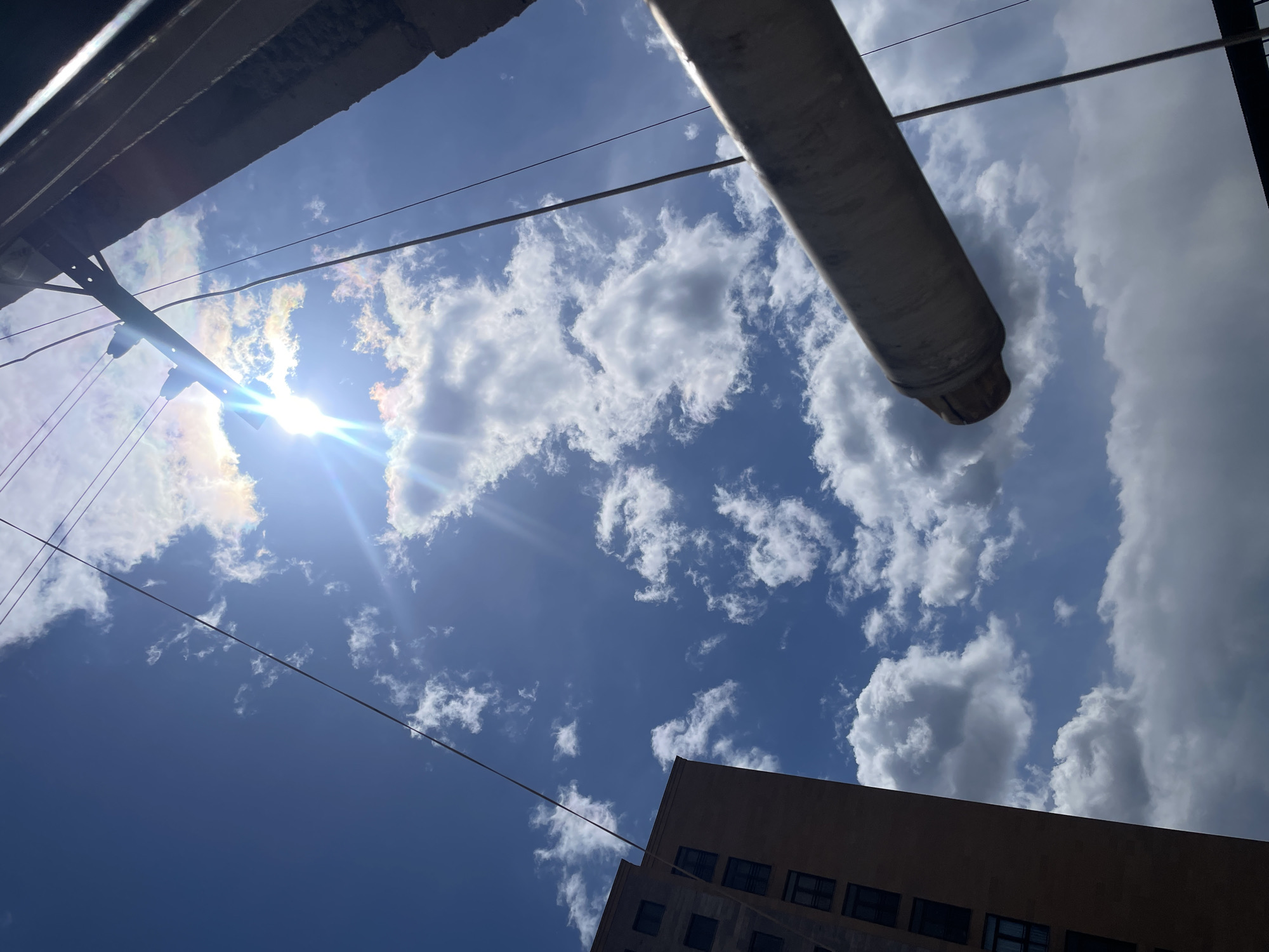 The image is a low-angle shot of a sky in Yerevan, Armenia. The sky is blue with scattered cumulus clouds. The sun is visible, partially obscured by clouds, creating a bright glare. In the foreground, there is a building with windows and an overhanging structure made of what appears to be concrete. Also, there is a cylindrical object, possibly a pipe, protruding into the frame from the upper right corner. Power lines are also visible traversing the sky. No people are visible.