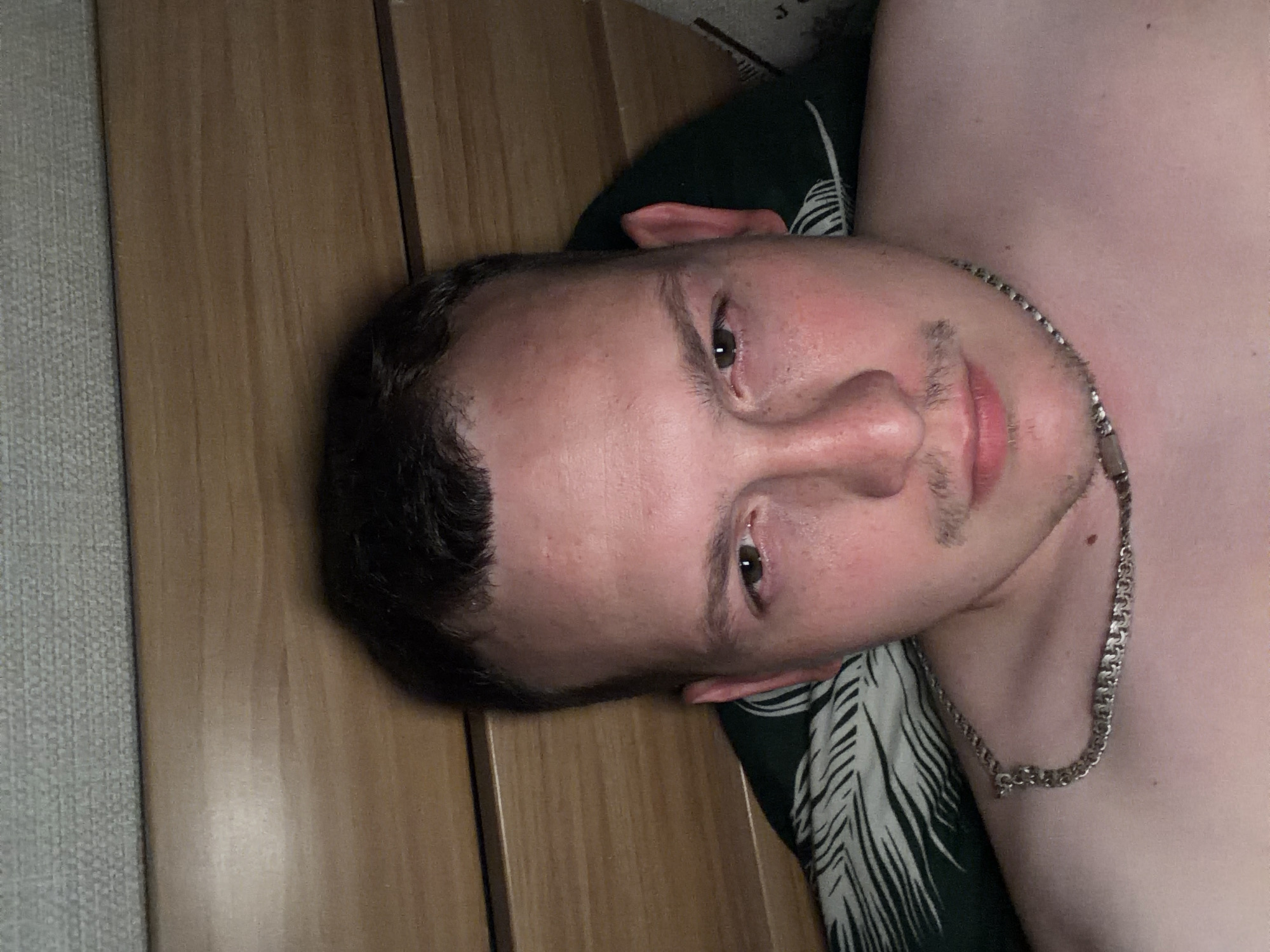 A man is lying in bed, looking at the camera. He has short brown hair, a mustache, and is wearing a silver chain. The background is a wooden headboard and a pillow with a leaf pattern. The location is unknown, but the image is from Chaikovsky, Russia.