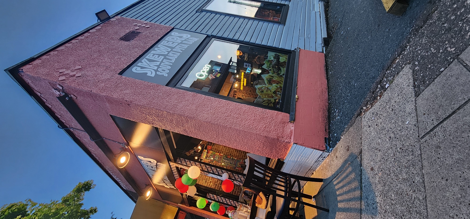 The image shows the exterior of a restaurant named "SKEWERS SOUVLAKI PITA BAR" in an urban setting. The building has a textured pink stucco facade on one side and a gray horizontal siding on another. There are large display windows showcasing the interior, which appears to be a dining area with brick walls. Inside, there are images of food items, and a neon "Open" sign is visible. Outside, a sidewalk with a concrete texture runs along the front of the building, and a black metal chair is placed near the entrance. A string of red, green, and white balloons is attached to the building's exterior. The lighting suggests it is either dawn or dusk. The sky is a clear, deep blue.
