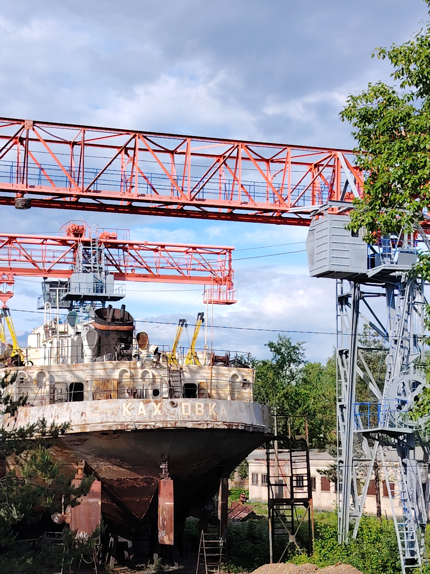The image depicts a portion of the rusting hull of a ship named "Kakhovka" in Chaikovsky, Russia. The ship is grounded, not in water. A complex system of red overhead cranes is positioned above the ship. To the right, a tall, gray crane structure stands amidst green foliage. The sky above is partly cloudy. Various ladders and support structures surround the hull. Buildings are visible in the background.