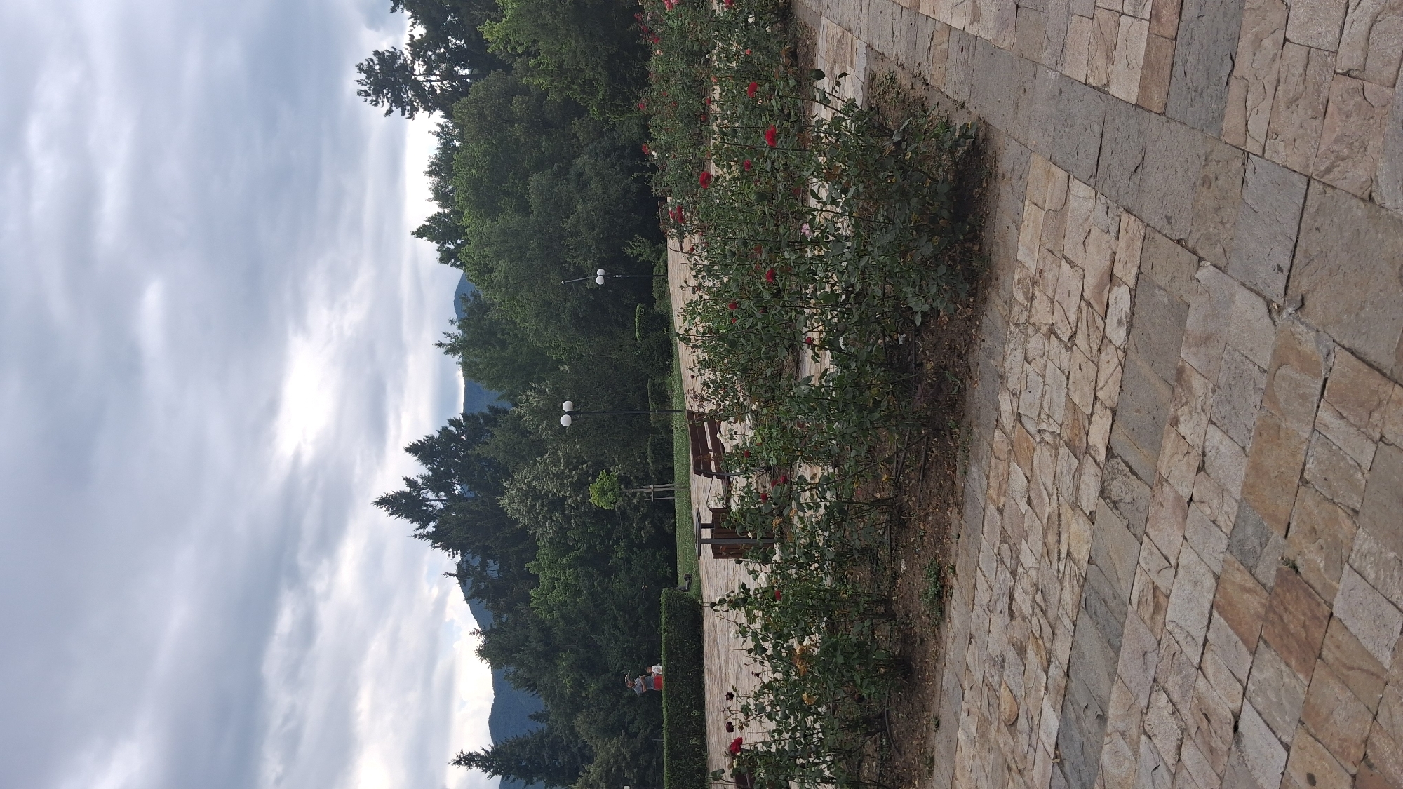 The image depicts a park area with a paved walkway made of irregular stone slabs. A rose garden with numerous red and pink roses is visible in the foreground and midground. In the background, there are trees and a cloudy sky. A person is standing near a hedge, and a wooden bench and a small table are placed on the walkway. This scene is located in the city of Gotse Delchev, Bulgaria.