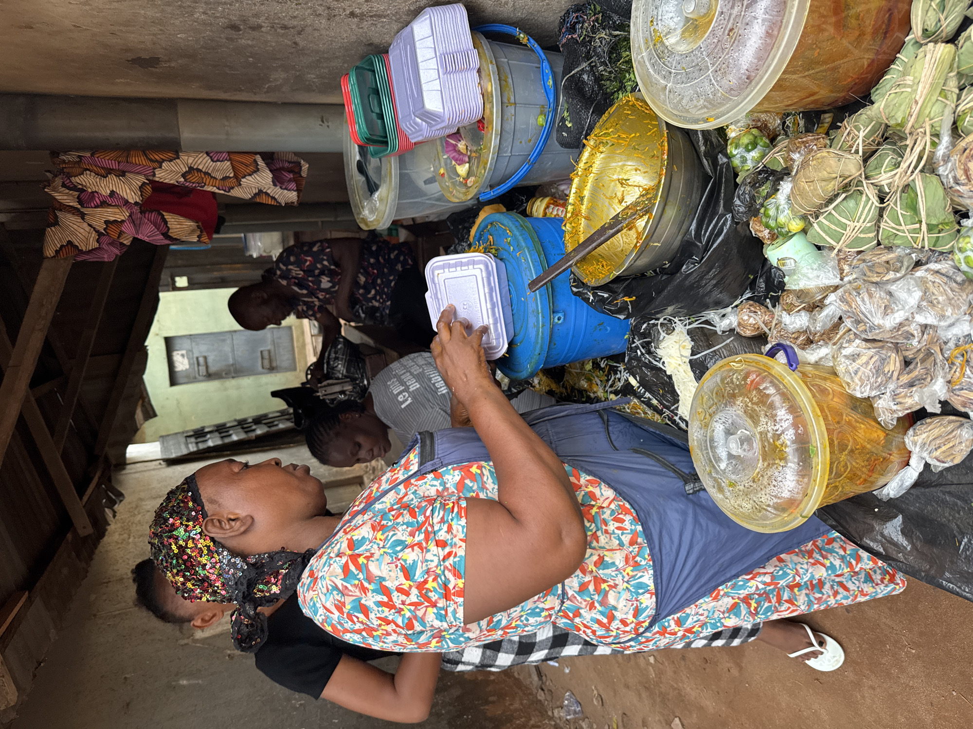 A woman stands in a street market in Onitsha, Nigeria. She wears a colorful headscarf and dress, and white flip-flops. To her right are several buckets and containers holding food items, including bundles wrapped in leaves. Behind her are other people, and a building is visible in the background.