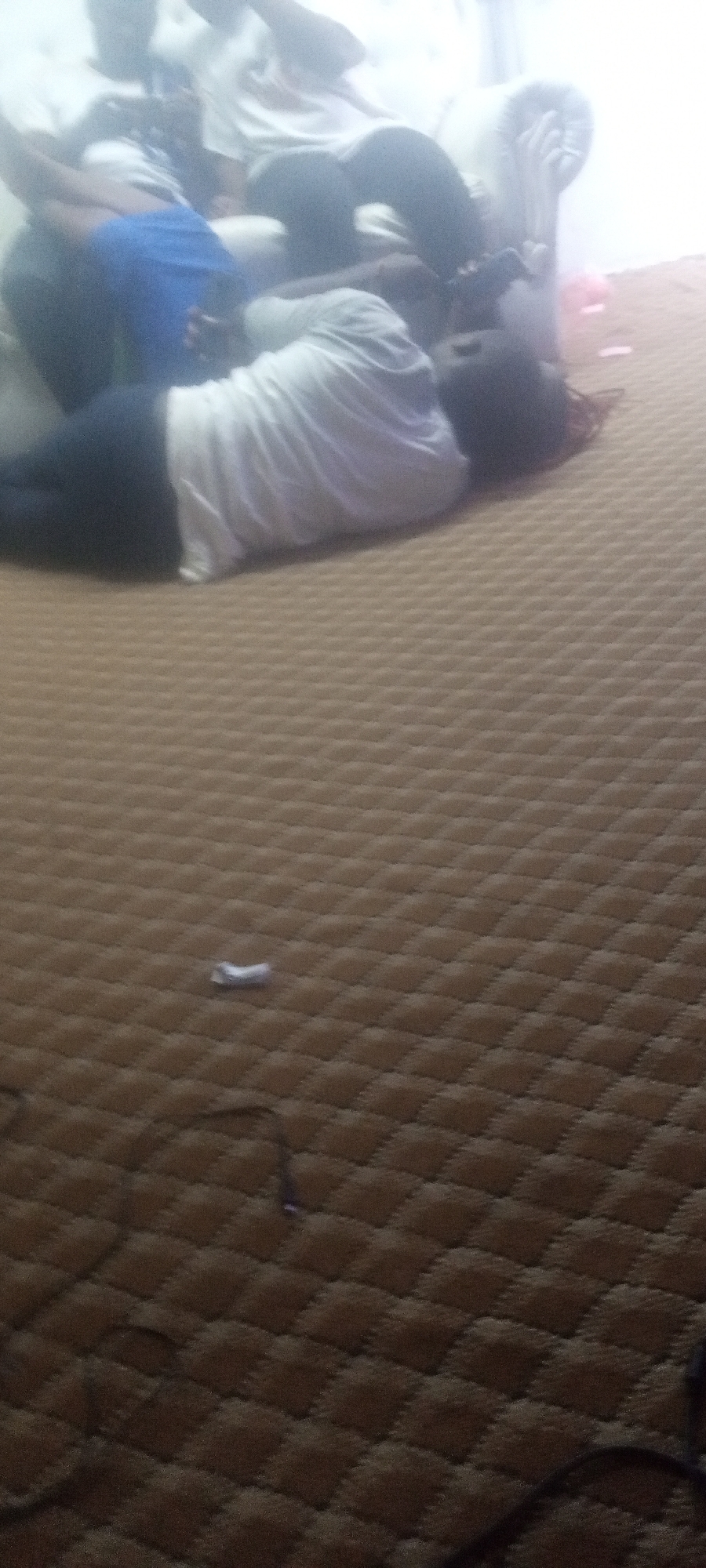 A poorly lit interior depicts several individuals. Some are seated on a white couch, while one person lies on the carpeted floor. An unidentified object lies near the prone figure. The location appears to be an interior setting in the city of Unknown, Unknown. Additionally, a black wire or cable is visible extending across the carpet in the foreground of the image.