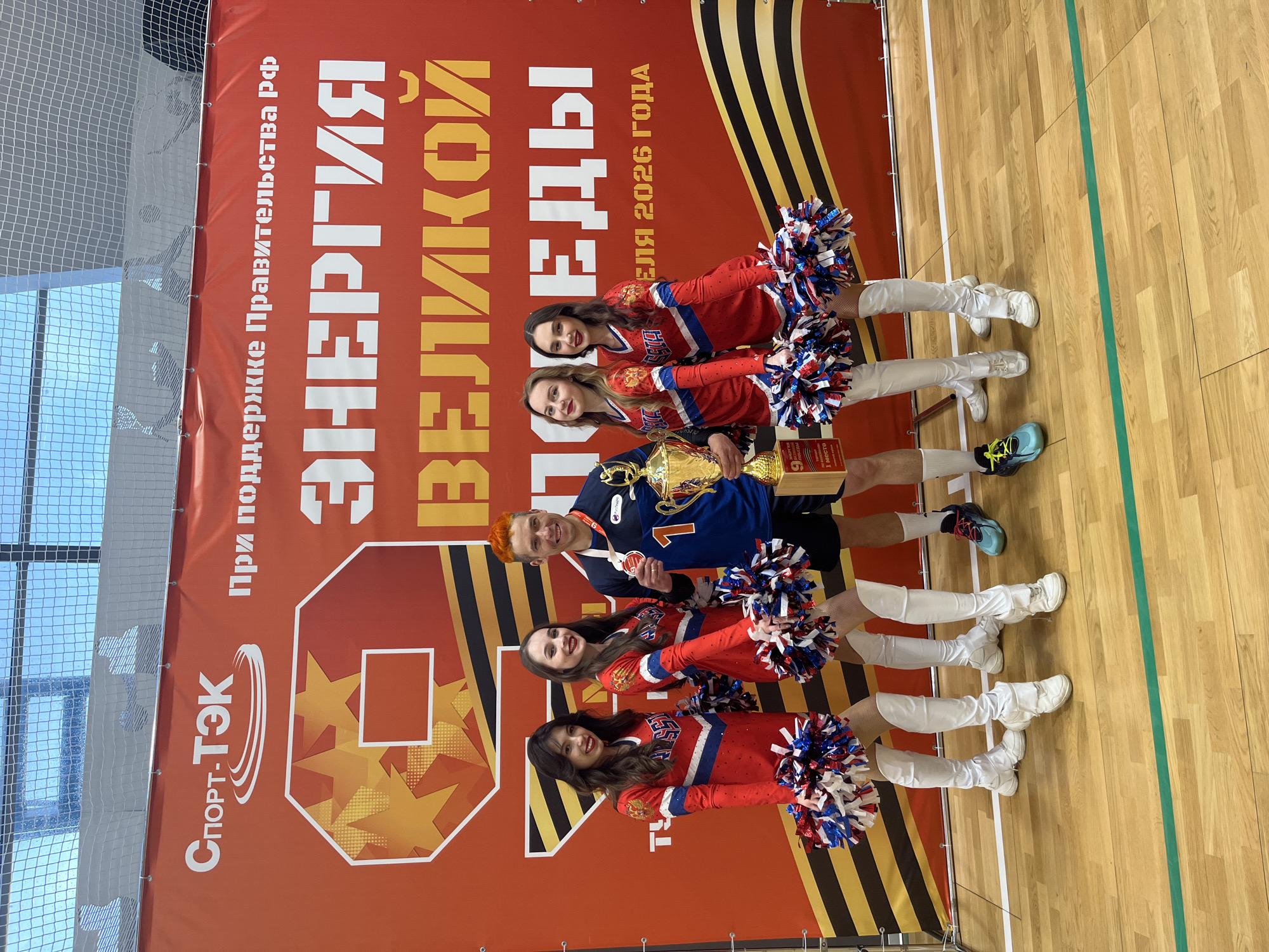 Here is a detailed description of the image:
**Scene:** The image appears to be taken indoors, possibly in a sports hall or gymnasium. A group of people are posing in front of a large banner.
**Banner Details:** The banner is predominantly red and features white text in Russian. The text includes phrases like "Under the support of the Government of the Russian Federation," "Energy," "Great," and "Victory." The text includes the year 2026. A stylized "9" is visible on the banner, decorated with yellow stars. The logo "Sport-TEK" is present in the bottom left corner.
**People:** The image includes five individuals. One male, wearing a blue jersey with the number "1" on it and an orange hair color, holds a golden trophy. He also has a medal around his neck. Surrounding him are four females, all dressed in matching red and white cheerleading outfits with white boots and holding pom-poms in red, white, and blue colors.
**Objects:**
* A golden trophy.
* Pom-poms in the cheerleaders' hands.
* A medal around the male's neck.
**Environment:** The floor of the location is made of wood, with white and green lines painted on it. In the background, a netted fence is visible on the left.
**Based on the Russian text and the symbolism on the banner (such as the stylized "9" with stars, reminiscent of Victory Day celebrations), this is likely a scene related to a sports event or competition in Russia, possibly in Moscow.**