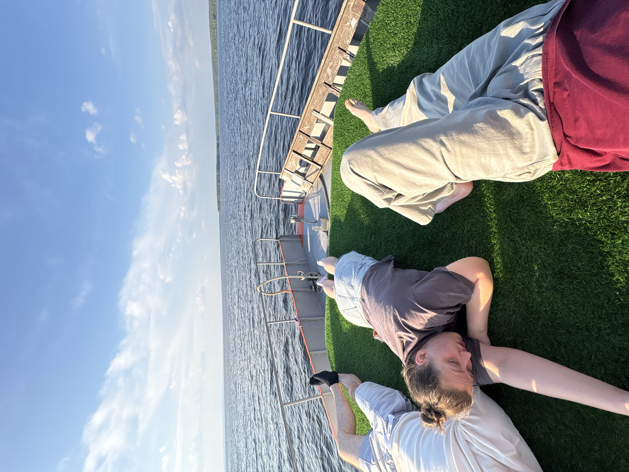 Four individuals are lying on the deck of a boat, which is covered in artificial grass. The boat is on a body of water, with a treeline visible in the distance. The sky is blue with scattered clouds. The individuals appear to be relaxing or sleeping. One person is wearing light-colored pants and a burgundy shirt. Another person is wearing light-colored pants and a dark shirt. The third person is wearing denim shorts and a dark shirt. The fourth person is wearing light-colored shorts and a black sock on one foot. The location is the Kama River in Chaikovsky, Russia.