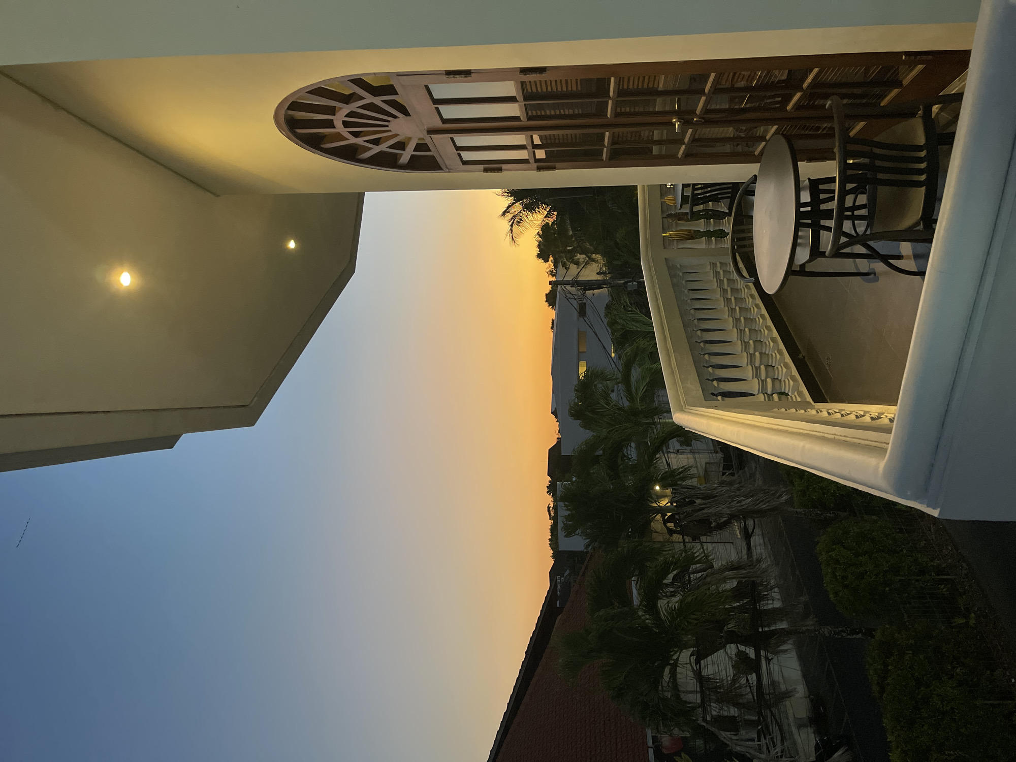 The image shows a view from a balcony in North Kuta, Indonesia, at dusk or dawn. The sky transitions from blue to orange. In the foreground, there's a table and chairs on the balcony, framed by a white balustrade. A building's architecture is visible, featuring an arched window or doorway with a wooden frame. Below the balcony are trees, rooftops, and parts of buildings, suggesting an urban or suburban environment.