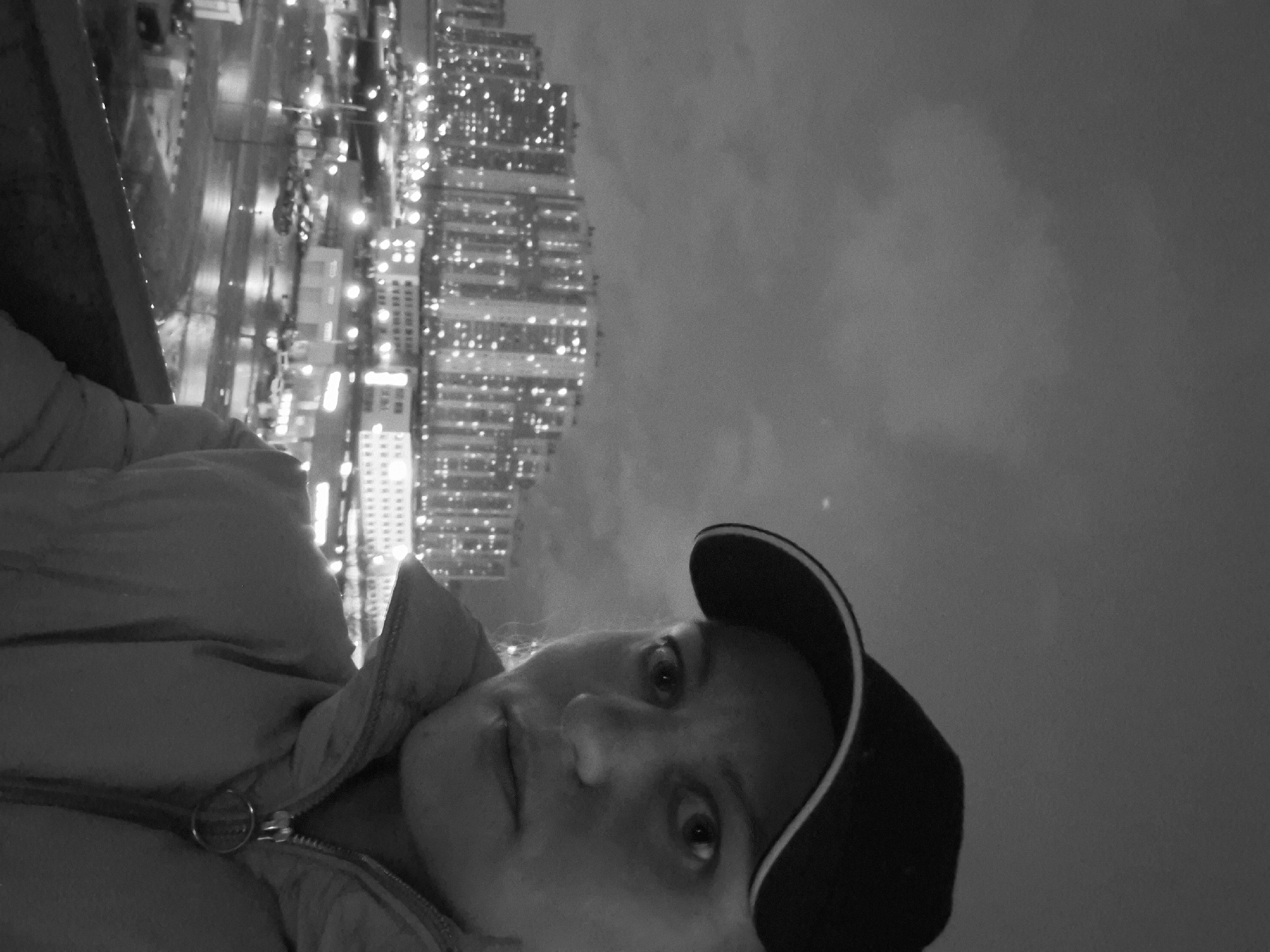 A grayscale, low-angle photograph captures a person with light skin wearing a dark baseball cap and a puffy jacket. The person is positioned towards the lower right of the frame and looks directly at the camera. In the background, the city of Saint Petersburg, Russia, is visible. The cityscape is illuminated with numerous lights, suggesting it is nighttime. The upper right portion of the photo is dominated by a cloudy sky. The person appears to be on a high balcony or rooftop, overlooking the urban landscape.
