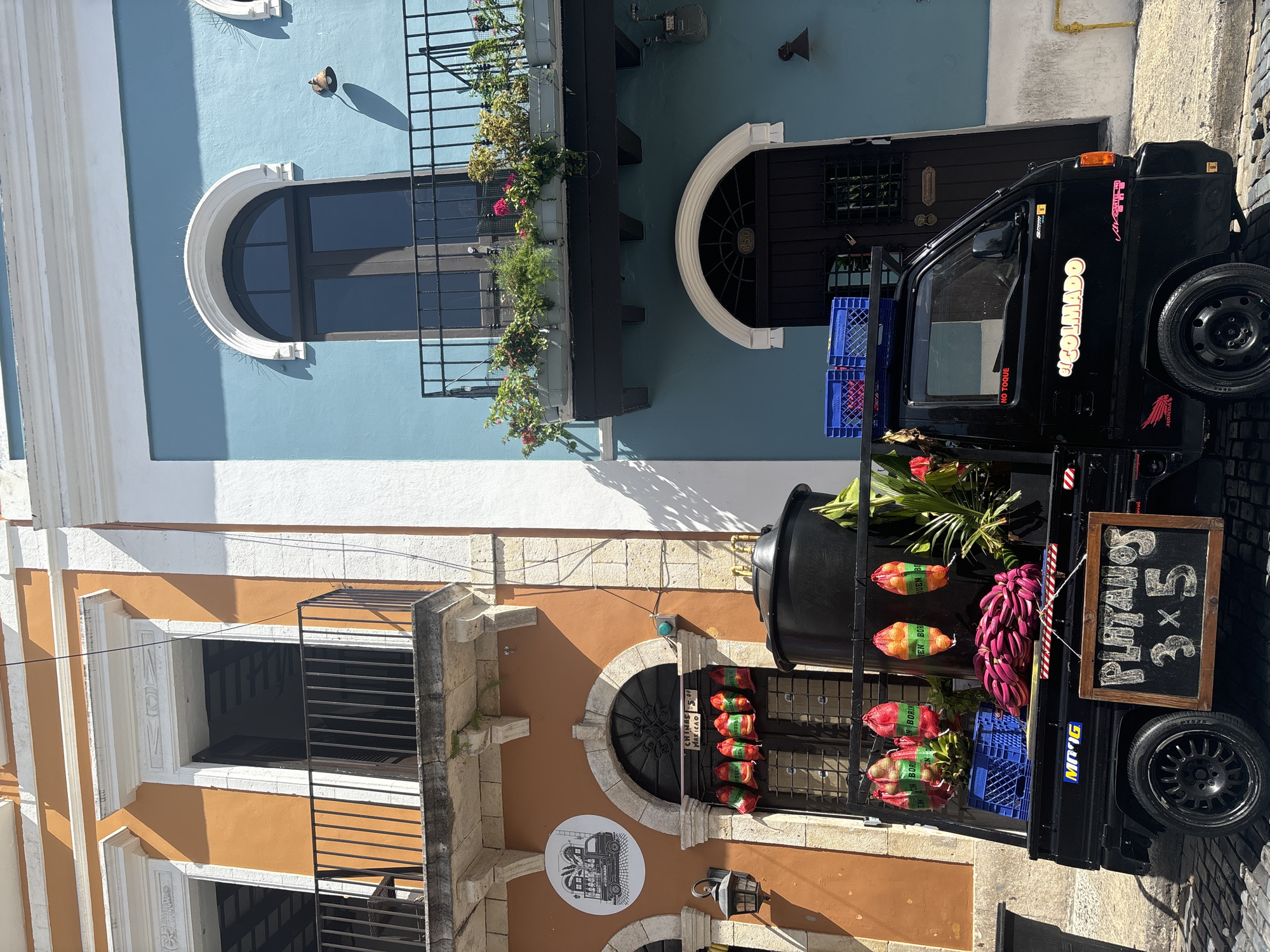 The image depicts a street scene in Old San Juan, Puerto Rico, United States of America, focusing on the colorful architecture and a parked black flatbed truck. The truck is laden with produce, including bananas and what appear to be plantains, some bagged and hanging, and others in blue crates. A chalk-written sign on the truck reads "PLATANOS 3x5". To the left, an orange building features a round sign with a black and white illustration of a truck. Red and green bags are hanging above a doorway on this building. To the right, a striking blue building rises, with an arched window and a balcony overflowing with potted plants. Both buildings exhibit architectural details common to Old San Juan, such as balconies with black metal railings and ornate window frames. The ground is paved with cobblestones, and a gas meter is visible on the exterior of the blue building. The scene is illuminated by bright sunlight, casting distinct shadows. There are no people clearly visible in the frame.
