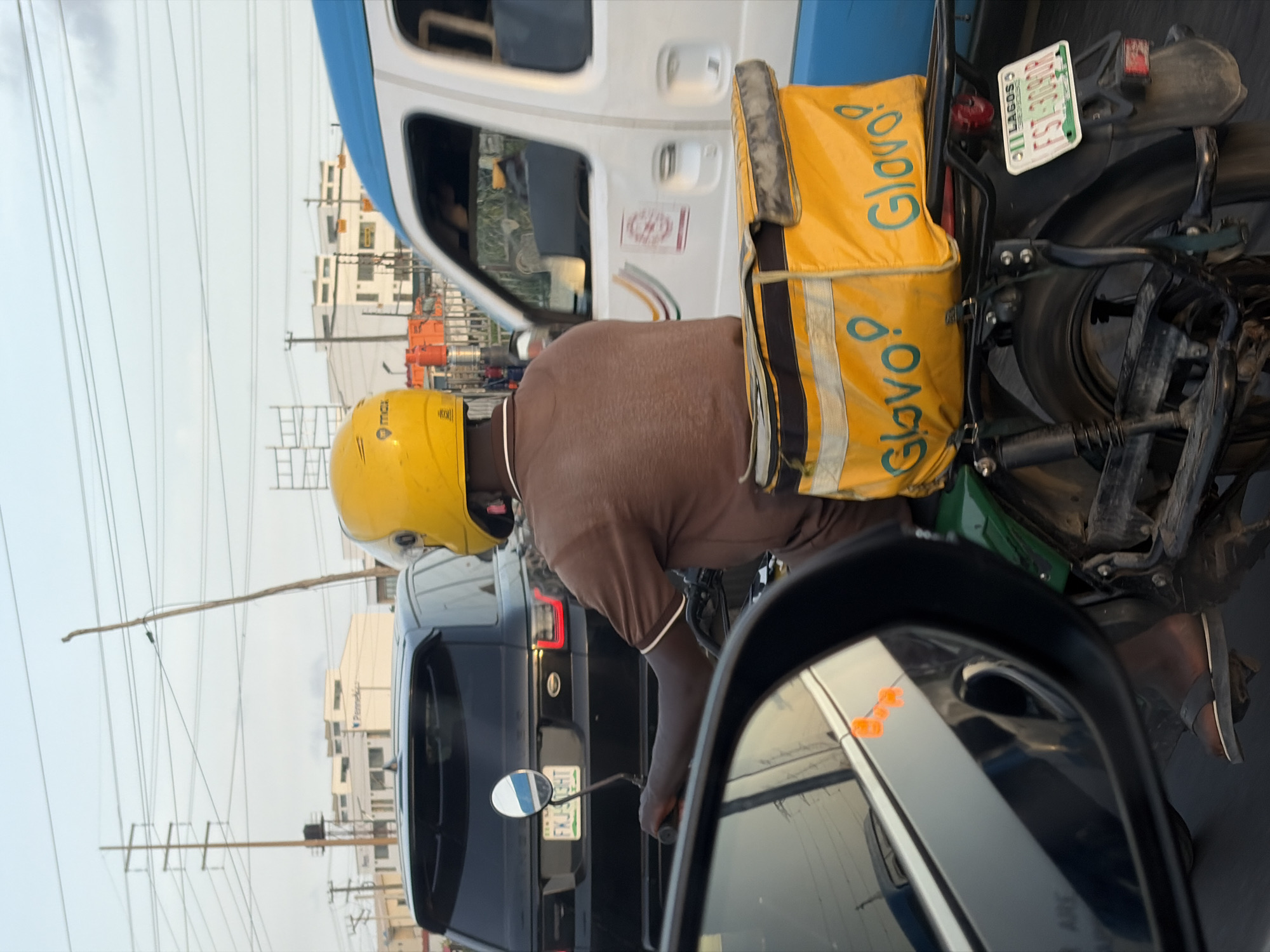 A Glovo delivery driver is on a motorcycle in traffic in Igboefon, Nigeria. The driver is wearing a yellow helmet and a brown shirt. The motorcycle has a green frame and a yellow Glovo delivery bag attached to the back. The license plate on the motorcycle reads "11 LAGOS FST309GR". A black SUV is visible in the background, its license plate partially obscured but appearing to read "EKJ309HT." A white van with blue accents is also present. The scene is captured through a car's side mirror, reflecting orange lights from other vehicles. Power lines stretch across the sky.