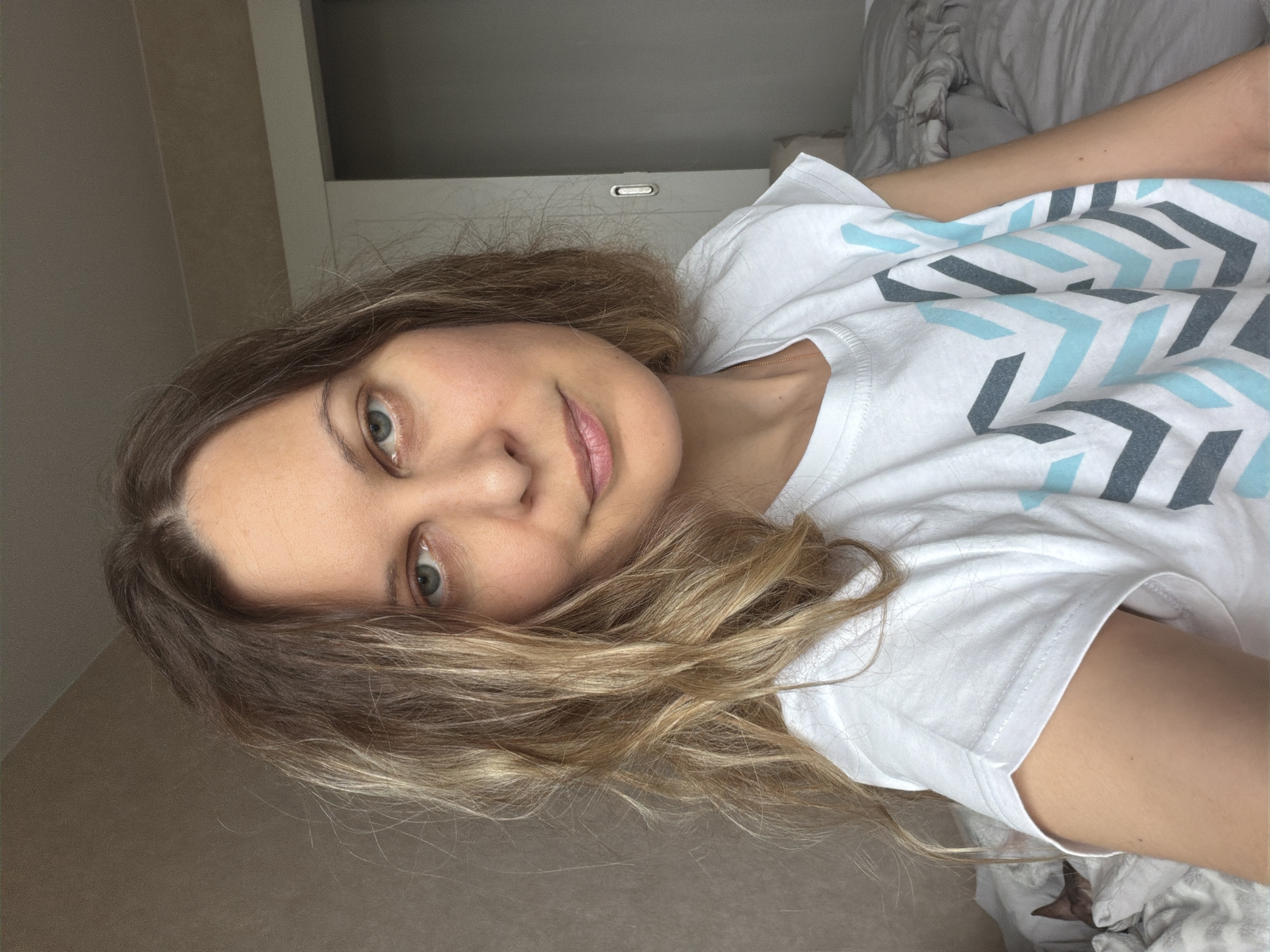 A woman with shoulder-length, blonde-highlighted hair, blue eyes, and fair skin is lying down, facing the camera. She is wearing a white T-shirt with a geometric pattern of light blue and gray chevrons. The background consists of a beige-colored wall and a white wardrobe cabinet. The scene appears to be indoors, possibly in a bedroom, with part of a rumpled bed visible in the upper right corner. Location is Unknown, Unknown.