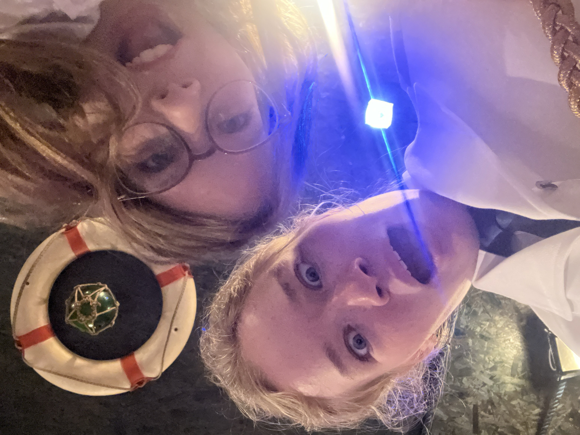 Two light-skinned women are photographed from a low angle, looking down at the camera with exaggerated facial expressions: one with glasses and the other with her mouth open. To the left of the frame is a decorative life preserver with red bands, positioned on a dark surface. Bright, diffuse light illuminates the subjects, with an additional point light source casting blue streaks in the image center. The woman on the right wears a white collared shirt with gold braiding at the shoulder. The scene appears to be indoors, possibly a restaurant or bar, suggested by the patterned surface at the bottom right of the frame. The location may be in the city of Budva, Montenegro, although no explicit location details are visible in the image.