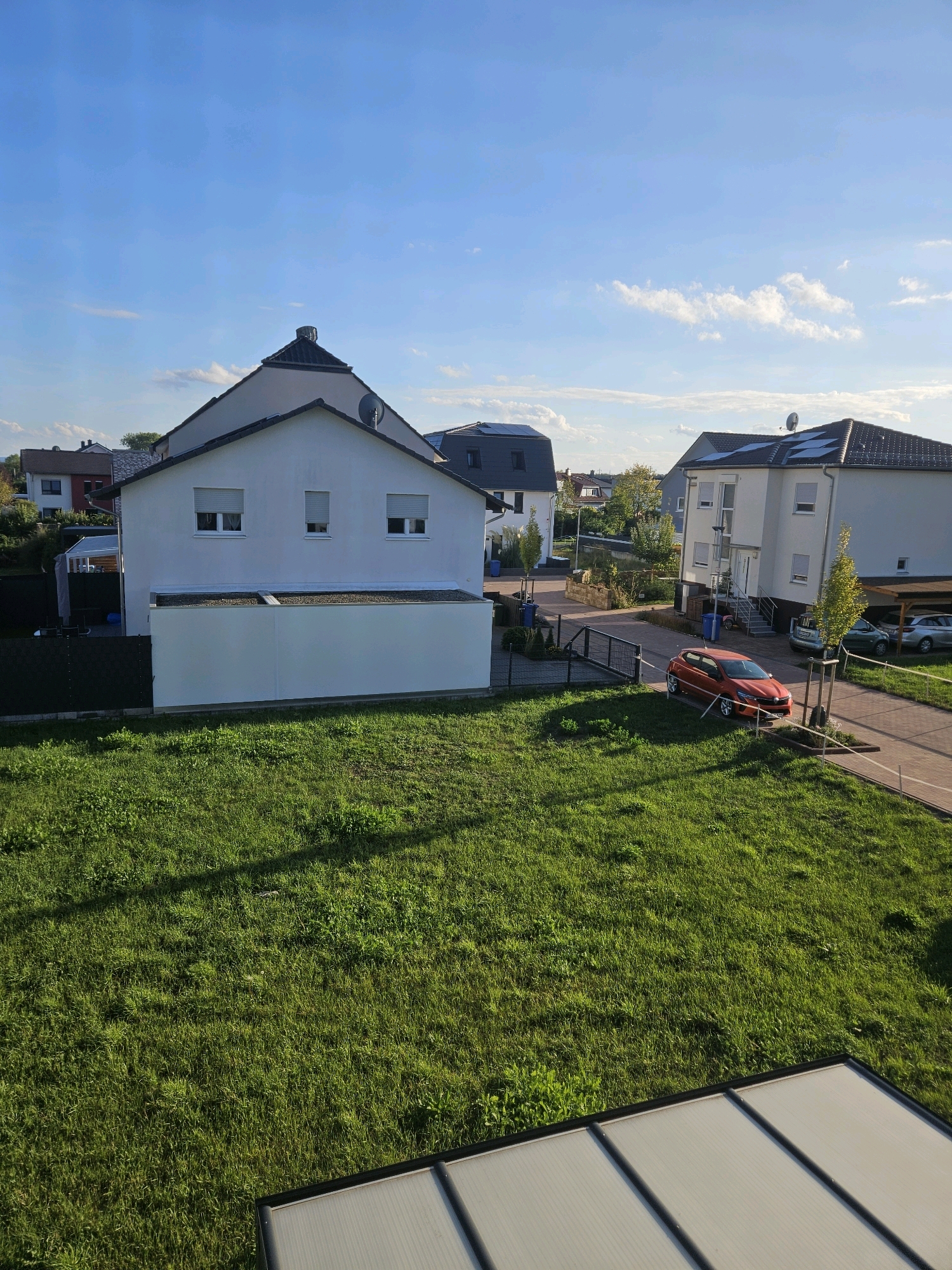 The image depicts a suburban street scene in Eppertshausen, Germany, under a clear blue sky with scattered white clouds. In the foreground, a lush green lawn is visible, with a shadow cast across it. Beyond the lawn, a row of modern residential buildings lines the street.
To the left, a two-story white house with a dark sloped roof and three windows on the upper floor is prominent. A white wall with a dark fence runs along the front of this property. To the right of this house, a narrower, darker-colored building with a sloped roof is partially visible.
Further down the street, a larger, white, three-story building with multiple windows and a dark sloped roof stands to the right. A driveway leads to this building, with a covered carport to its right. An orange hatchback car is parked on the driveway. In front of the buildings, there are trees and bushes. Blue recycling bins are placed near the entrance of the larger building on the right. The road surface appears to be paved with bricks.
The overall impression is of a well-maintained residential area on a sunny day.