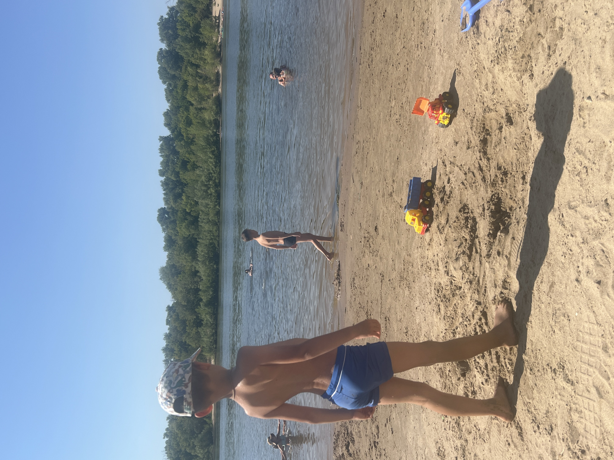 The image depicts a sunny day at a river beach, likely in Kilyakovka, Russia, as indicated by the context. In the foreground, a young boy wearing a patterned cap and blue swim trunks stands facing away from the camera, his back to the river. His stance suggests he is observing something in the water or on the sand. The sandy beach is scattered with children's toys, including a yellow and blue toy truck and a red and yellow toy excavator. A blue toy shovel lies to the right.
In the middle ground, the river is populated with several people. A boy in dark swim trunks walks through the shallow water. Further out, a group of people, including at least one adult and a child, are swimming or wading. The river is calm with a clear blue sky overhead.
The background features a dense line of green trees along the far bank of the river, indicative of a natural, possibly wooded, area. The overall scene conveys a relaxed, recreational atmosphere typical of a summer day at a riverside location.