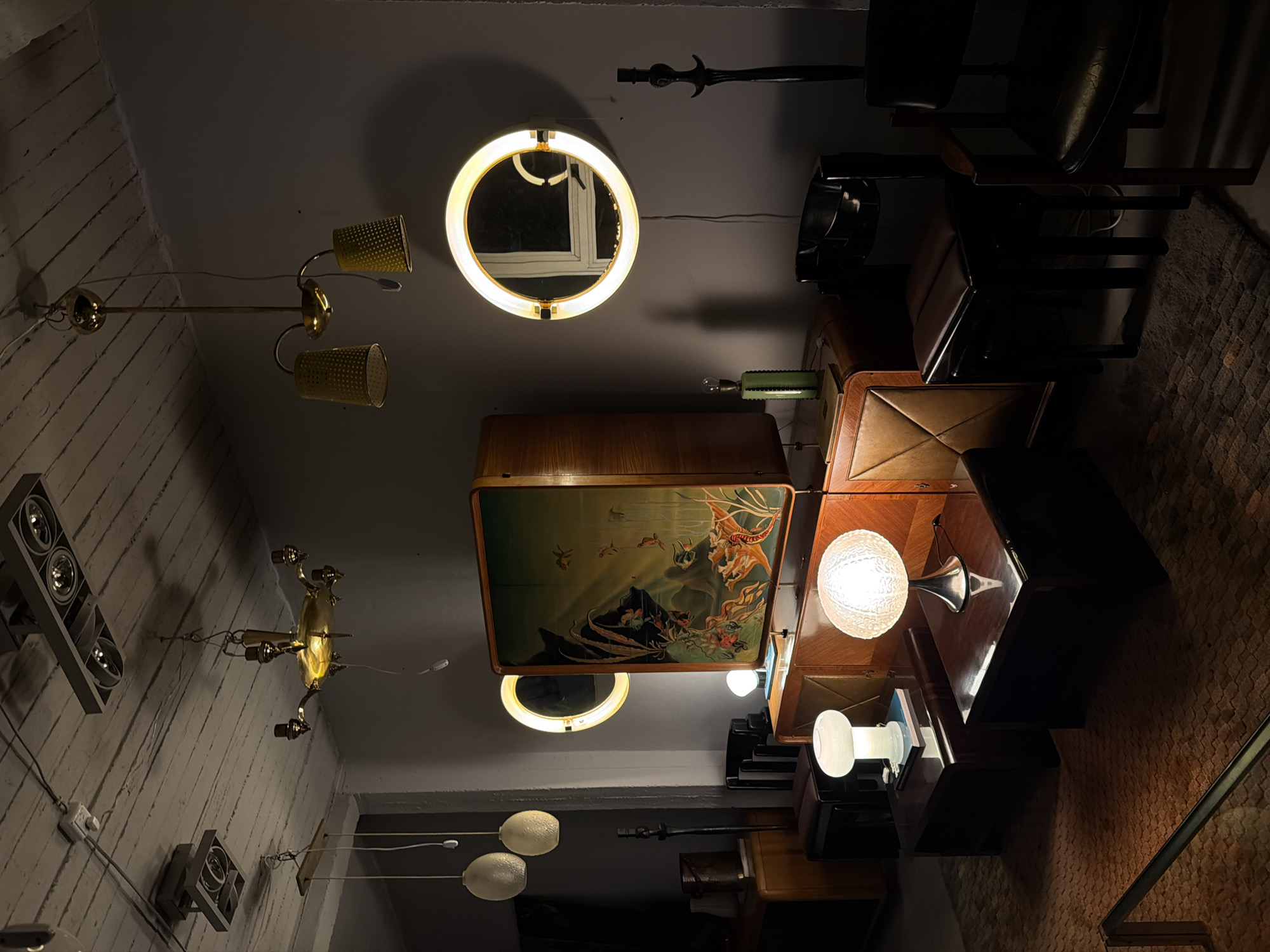 The image depicts an interior space in Buenos Aires, Argentina, filled with an eclectic collection of vintage furniture and lighting fixtures. The room is illuminated by various light sources, including a circular fluorescent lamp, multiple brass chandeliers with cone-shaped shades, and several table lamps, one with a globe-shaped shade. The walls are painted a muted grey, and some areas exhibit a textured surface. Several mid-century modern chairs with leather upholstery are visible, along with wooden cabinets and tables. A prominent piece of furniture features a painted scene, possibly a stylized depiction of mythology or folklore. The composition is organized with a focus on the arrangement of the furniture and lighting elements, creating a dense and cluttered yet aesthetically curated scene.