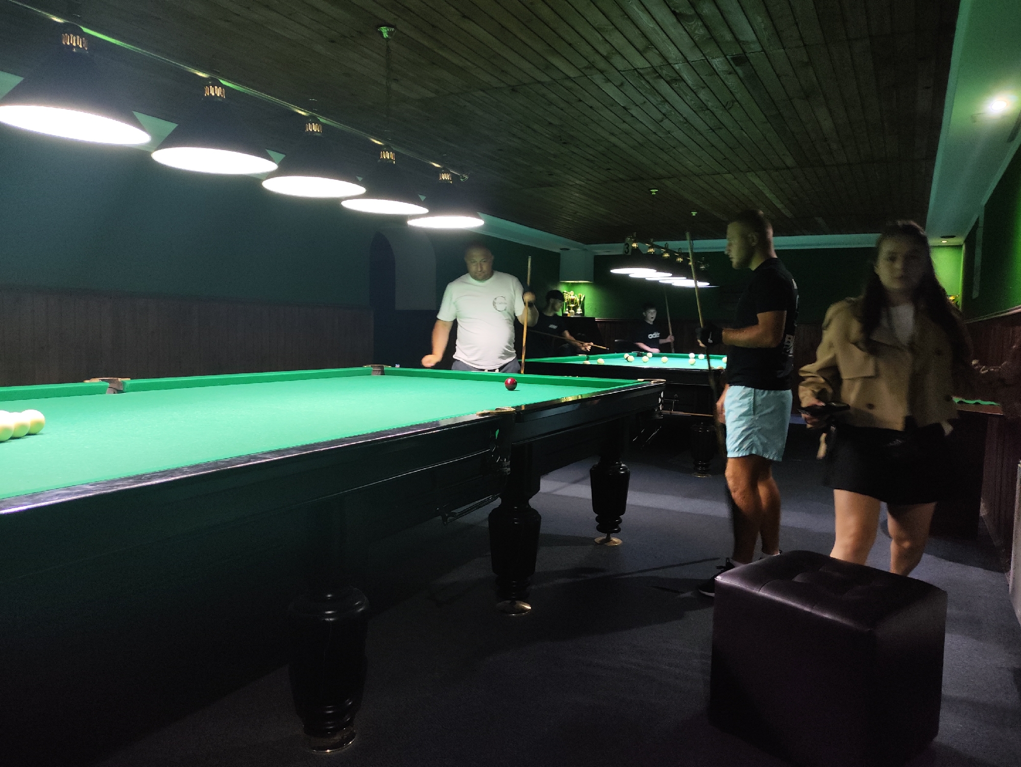 The image depicts an indoor billiards hall. In the foreground, a pool table with green felt and a red ball is visible. Several white balls are arranged on the left side of the table. In the mid-ground, a man in a white t-shirt is holding a pool cue, seemingly in the process of playing. Further back, two other individuals are visible near another pool table, also holding cues. On the right side of the image, two people, a man in a black t-shirt and light blue shorts, and a woman in a beige jacket and dark skirt, are standing. The environment is dimly lit, with overhead lights illuminating the pool tables. The walls are painted green, and the ceiling is made of wooden planks. There are dark stools placed around the room. The scene suggests a recreational setting where people are engaged in a game of billiards. Based on available information, this location is identified as a billiards hall in the city of Strășeni, Moldova.