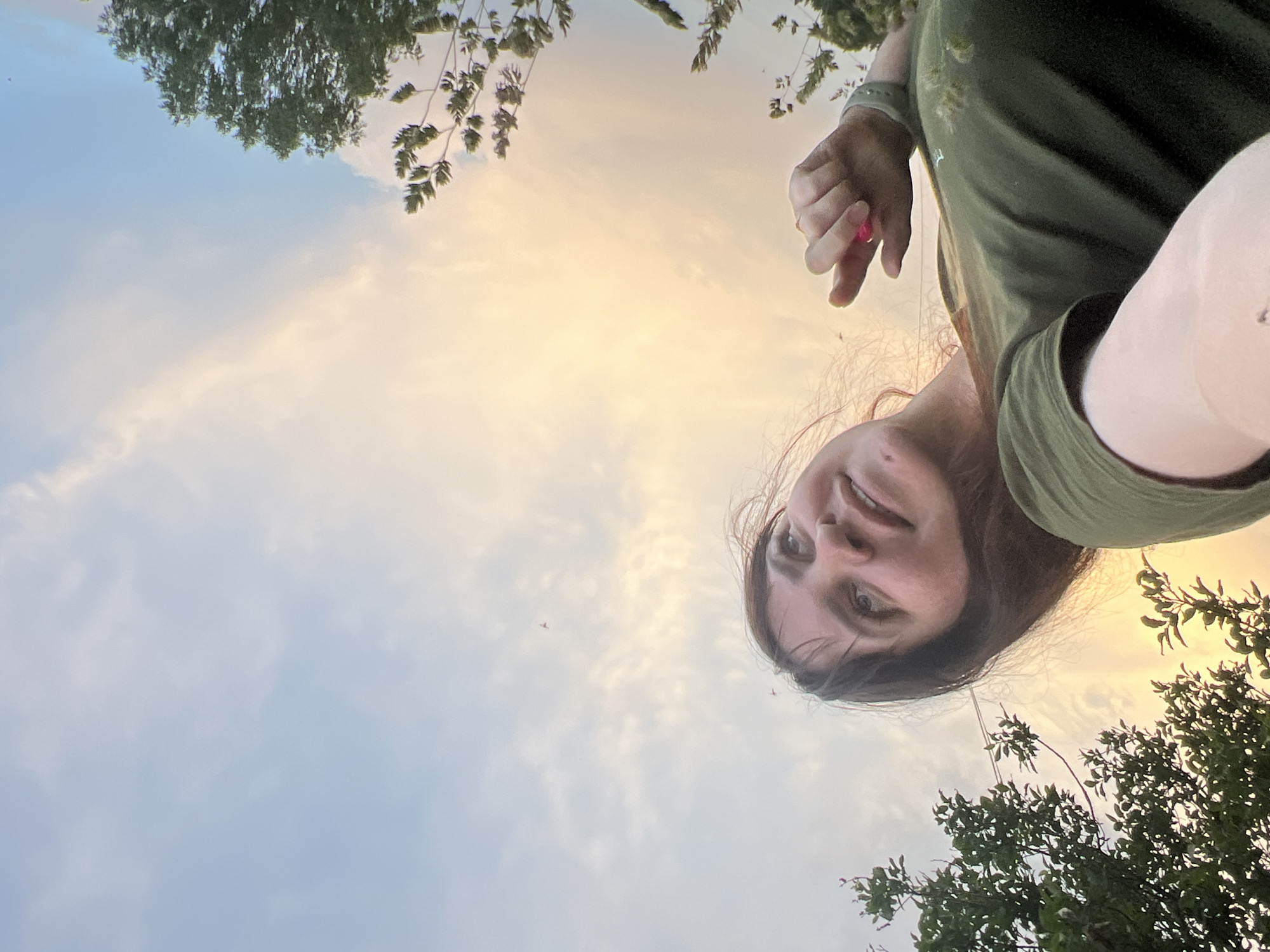 An outdoor, low-angle, medium shot captures a young woman with brown hair and bangs looking up at the sky. She is wearing a green t-shirt and has a pink object in her right hand, extended towards the camera. The background consists of a sky with scattered clouds, illuminated by warm sunset light, and foliage from trees framing the left and right sides of the image. The scene appears to be set in a natural or park-like environment within the city of Panteleevka, Russia.