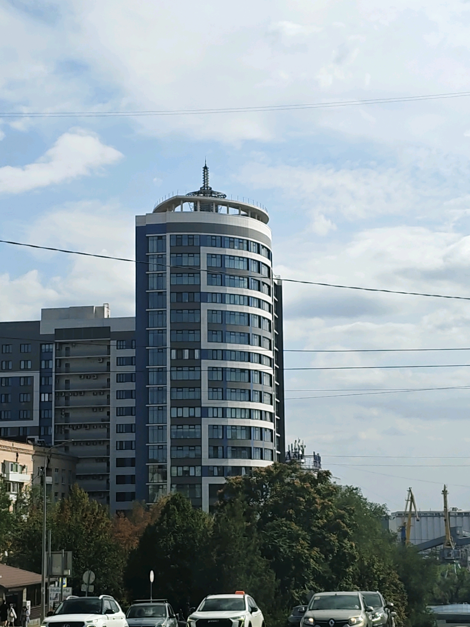The image shows a view of a city with a tall, curved building with blue and silver panels and numerous windows. To the left of this building is a taller, rectangular building with a similar color scheme and windows. The buildings appear to be in an urban setting, with trees and power lines visible. Several cars are present on a street in the foreground, and in the background, there are industrial structures with cranes, suggesting the presence of a port or industrial area. The sky is partly cloudy. The context of Rostov-on-Don, Russia is not visually apparent from the image itself.