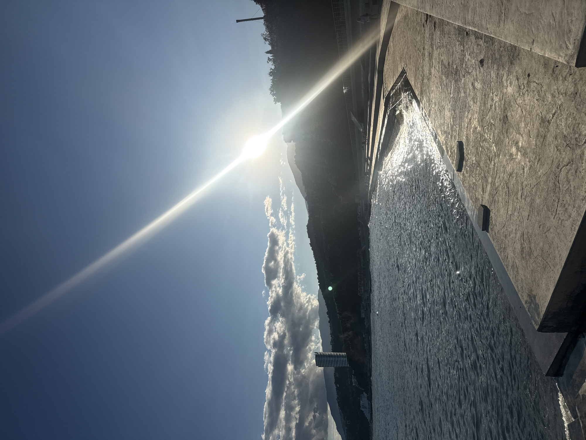 The image depicts a sun-drenched seaside promenade in Yalta, Ukraine. The sun, positioned high in the sky, casts a strong glare across the scene, creating a bright beam that stretches diagonally from the upper right to the lower left. The sea, a dark, rippling blue, laps against a curved concrete walkway that slopes down to the water. The promenade is lined with concrete steps and appears to be recently constructed or renovated.
In the distance, along the coastline, a prominent tall, slender building with a white facade stands out against the backdrop of lush, green hills. The hills are dotted with trees and more buildings, suggesting a developed urban area nestled into the natural landscape. The sky is a clear, bright blue, with a few scattered, soft white clouds that are catching the sunlight. The overall impression is one of a serene, sunny day at the seaside. There are a few people visible on the promenade, appearing as small, distant figures, engaged in walking or resting. The architecture and vegetation visible are consistent with a coastal resort town in southern Ukraine.