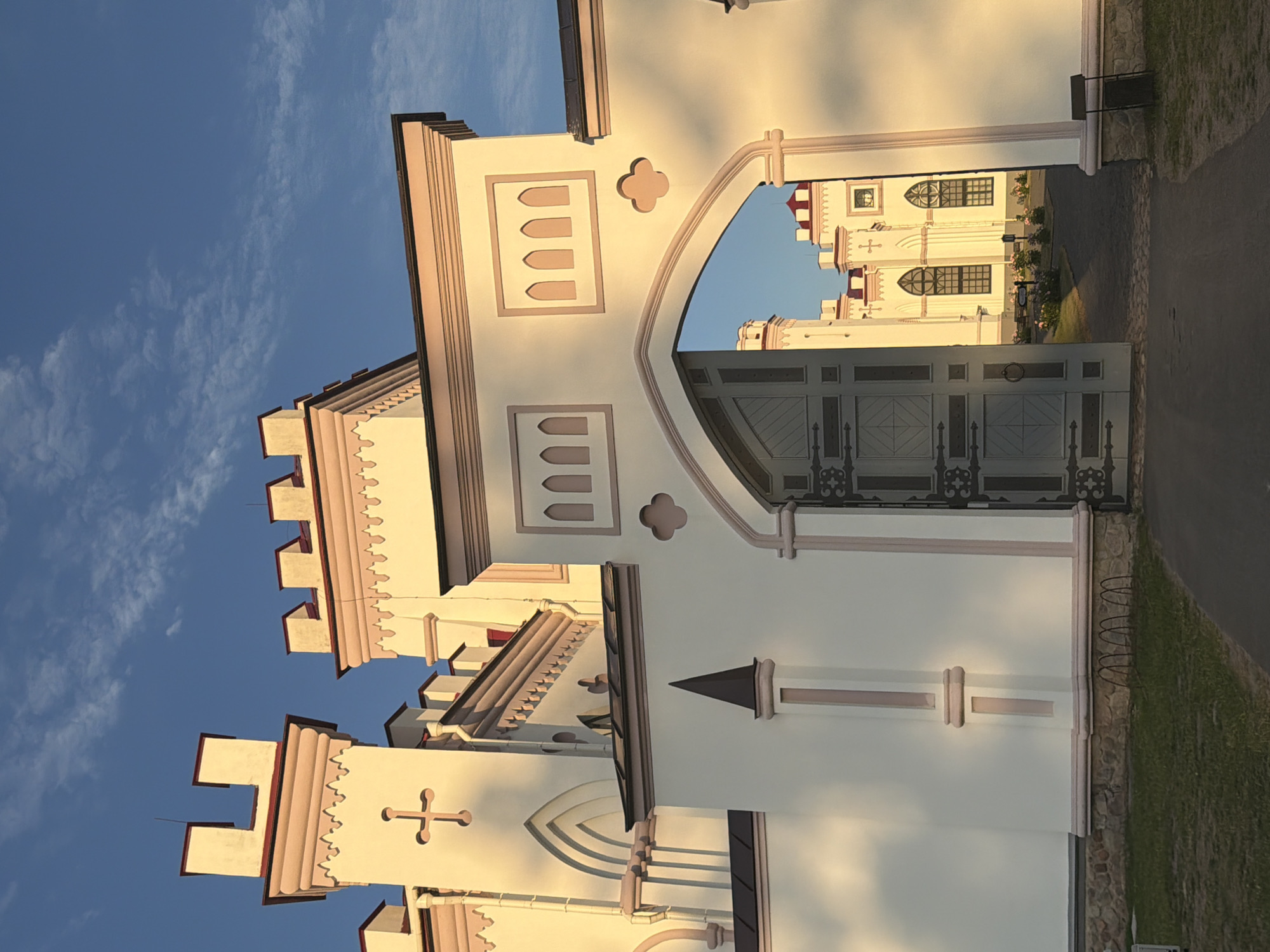 A light-colored, ornate castle with pale pink accents stands under a clear blue sky with scattered white clouds. The castle features crenellated towers, arched windows, and decorative molding. An open gate, made of dark metal with intricate scrollwork, leads into the castle courtyard. The sunlight casts long shadows across the facade, indicating it is either early morning or late afternoon. In the background, through the gate, more of the castle architecture and landscaping with some flowers is visible. The ground in front of the gate is covered with green grass.