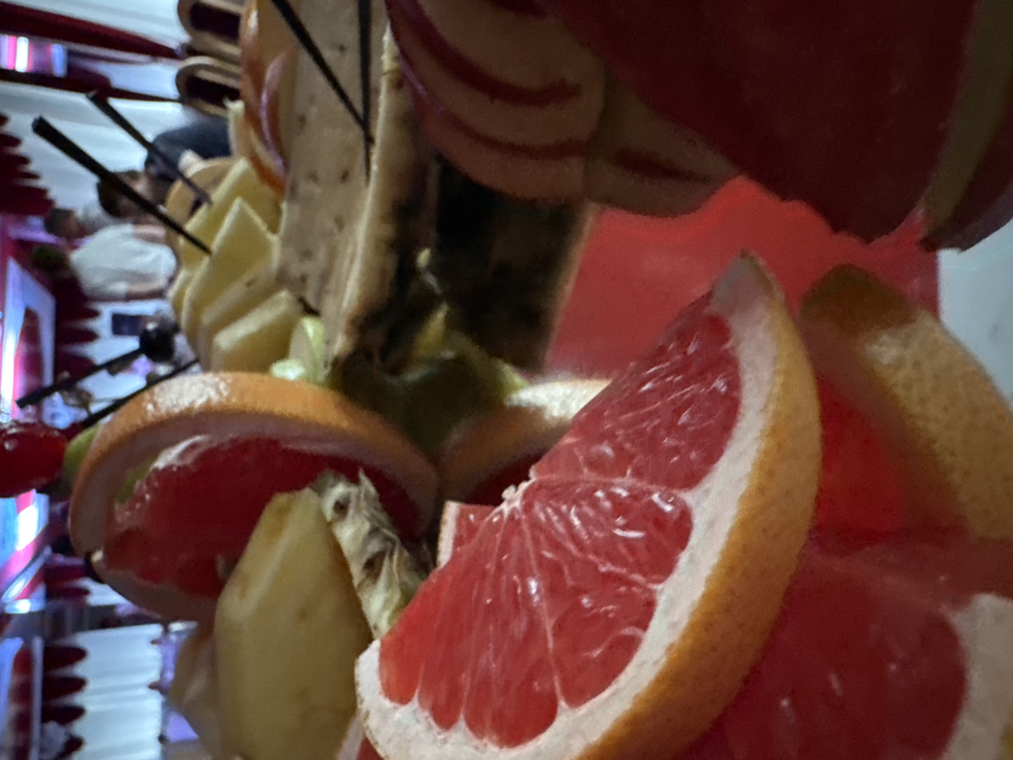 The image depicts a close-up, shallow depth of field view of a fruit platter featuring slices of grapefruit, pineapple, and possibly apple. The fruits are arranged on a reflective surface, likely a mirrored tray, with some appearing to be skewered with dark sticks. In the blurred background, a group of people are visible in what appears to be an indoor event space with red and white draped curtains and decorative elements. One person in the background is wearing a white shirt, and others are seated at tables. The overall ambiance suggests a celebratory or catered event. The image quality is moderate, with some blur and noise, particularly in the background elements. The specific location in Kryvyi Rih, Ukraine, is not discernible from the visual information provided.