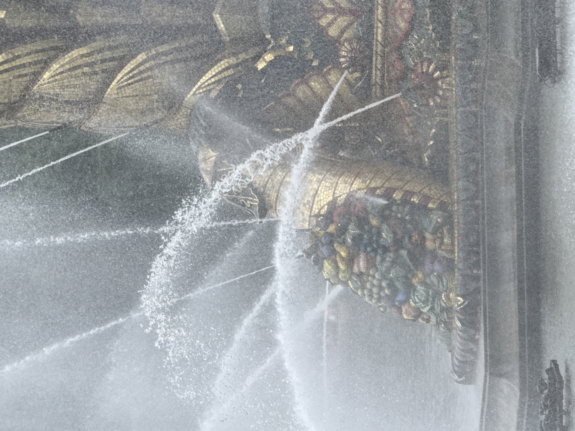 This image depicts a close-up view of a section of a fountain in Moscow, Russia, likely the iconic "Friendship of Peoples" fountain located at VDNH (Exhibition of Achievements of National Economy). The fountain features ornate mosaic work, with gold and dark tiles forming decorative patterns. In the foreground, a cluster of mosaic fruits, including grapes, pomegranates, and other colorful produce, is visible. Water is spraying upwards and outwards from various points of the fountain, creating a misty and dynamic effect. The base of the fountain is a tiered structure, also adorned with mosaic. In the lower left, there is a dark, low-lying object partially submerged in the water, which may be part of the fountain's structure or a decorative element. The overall scene is characterized by the play of water and light on the intricate mosaic surfaces of the fountain.