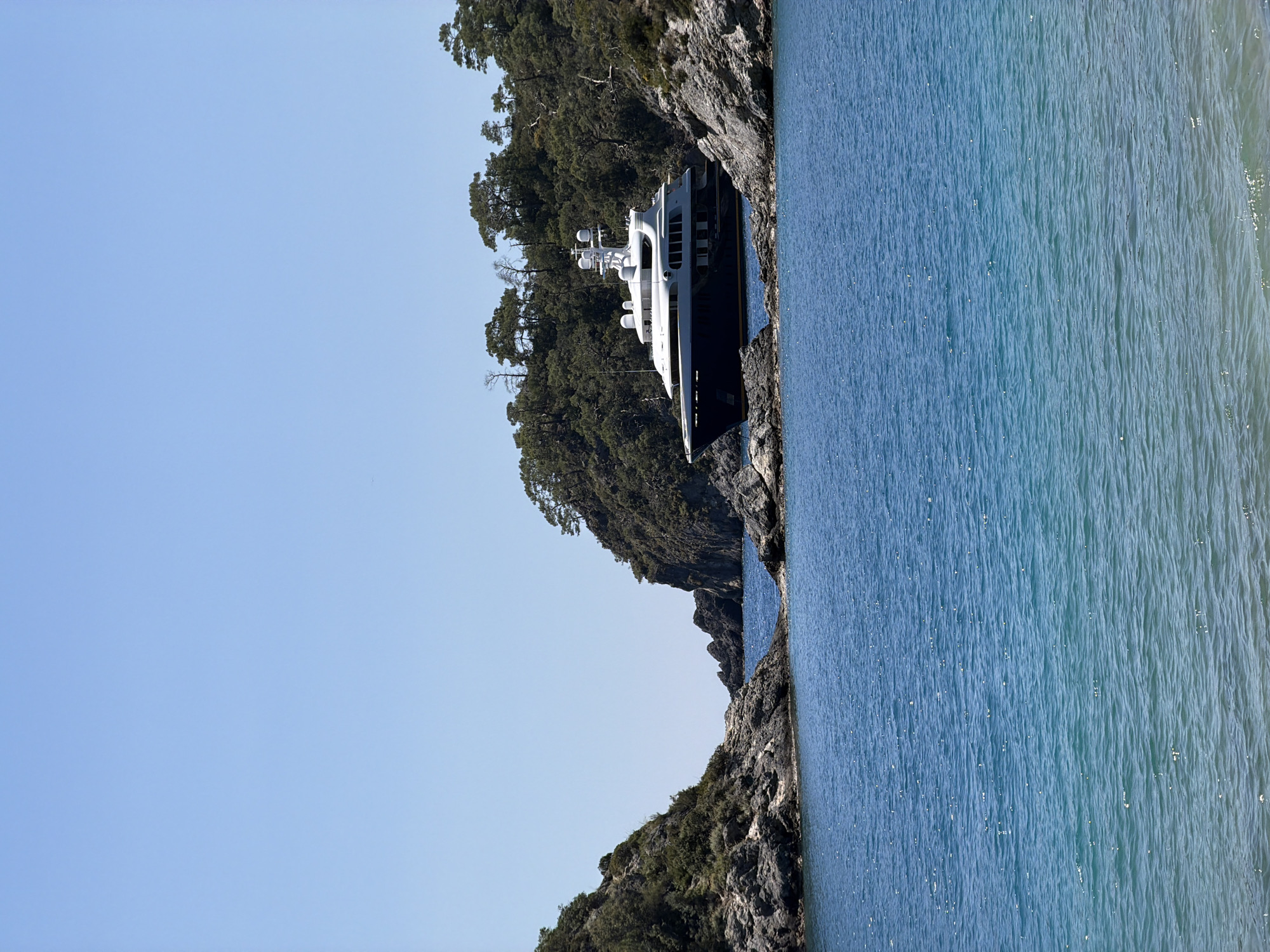 A sideways, eye-level shot depicts a large yacht nestled amidst rocky, tree-covered terrain along the coast, likely in the vicinity of Fethiye, Turkey. The yacht, predominantly white and black, appears docked or anchored close to the shore, almost integrated with the natural landscape. Its multi-level structure with visible antennas and satellite domes hints at luxury and advanced technology. The surrounding environment features jagged, rocky cliffs densely vegetated with trees. The water's surface adjacent to the boat reflects the sky and the trees, showcasing a blue hue with subtle wave patterns. The sky above is clear and light blue, indicating a sunny day.