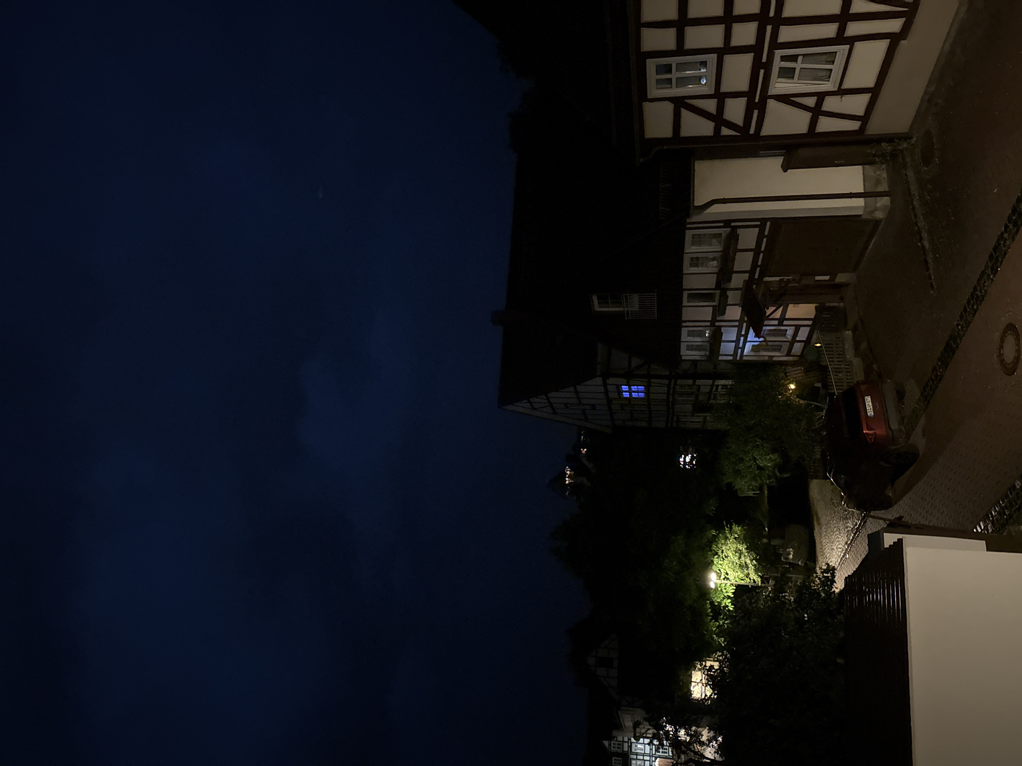 The image depicts a nighttime scene in a European-style town. The sky is a dark, cloudy blue, with a faint crescent moon visible. The foreground features a white-tiled roof. The middle ground shows a street paved with cobblestones, illuminated by a streetlamp. A red SUV is parked on the street. Several half-timbered houses with dark wood beams and light-colored infill are visible. Some of the windows in these houses are illuminated with a blue light. In the background, more trees and buildings are silhouetted against the night sky, with a distant tower or spire structure faintly lit.