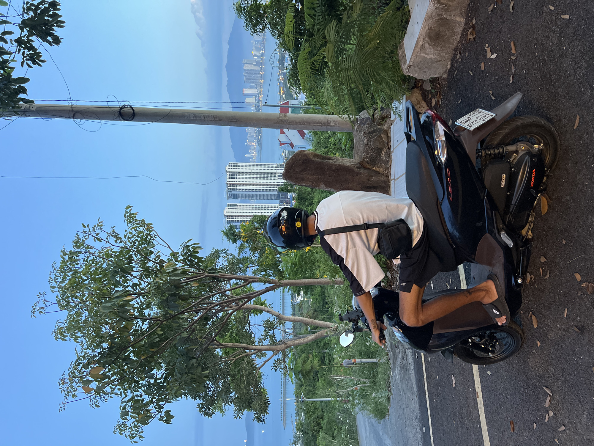 A person wearing a black helmet, white shirt with black sleeves, and black shorts rides a black scooter down a paved road. The person is facing away from the camera, looking towards a city skyline with tall buildings across a body of water. Trees and vegetation are visible in the foreground and background. A utility pole with wires stands to the right of the frame. The sky is a deep blue, suggesting dusk or early morning. The location is identified as Bac Nha Trang Ward, Vietnam.