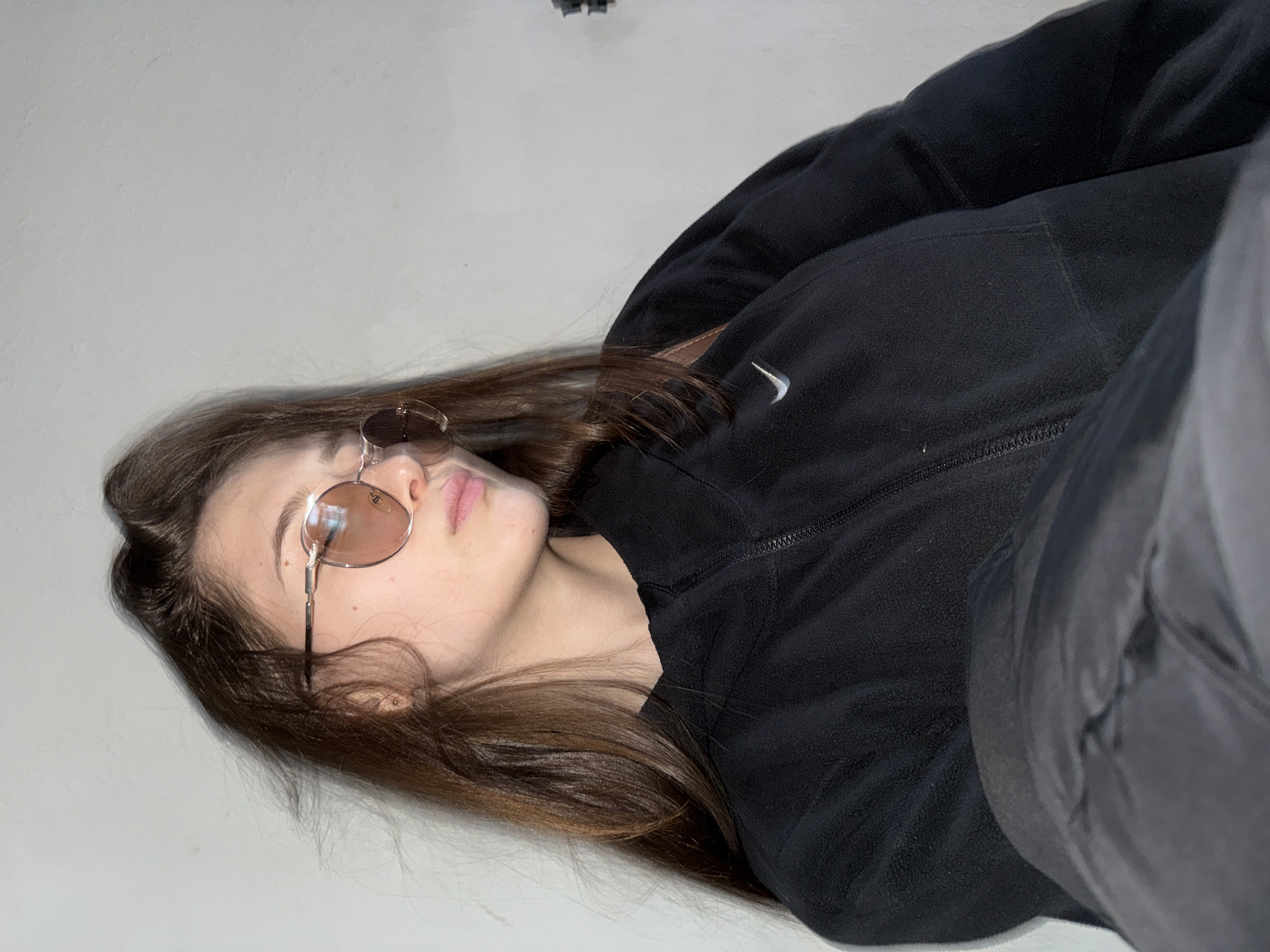 A young person, appearing to be female, is depicted in a tilted, close-up indoor shot, originating from Sofia, Bulgaria. She is lying down, with her long, dark brown hair spread around her head. Her face is turned slightly upwards, with her eyes concealed behind round-framed sunglasses that have a light amber or pink tint. Her lips are slightly parted, revealing a natural pink hue.
She is wearing a black zippered fleece jacket, clearly visible with a white Nike swoosh logo positioned on the left side of her chest. Part of a dark grey or black garment, possibly trousers, is visible on the right side of the frame. The background is a plain, light off-white or very light grey surface, suggesting a minimalist indoor setting without specific environmental cues. A small, dark, indistinct object can be seen at the top center of the background. The lighting is bright and even, indicating either diffused natural light or soft artificial illumination. The subject maintains a neutral, perhaps contemplative, expression, with the sunglasses adding a cool or reserved emotional tone to the image.