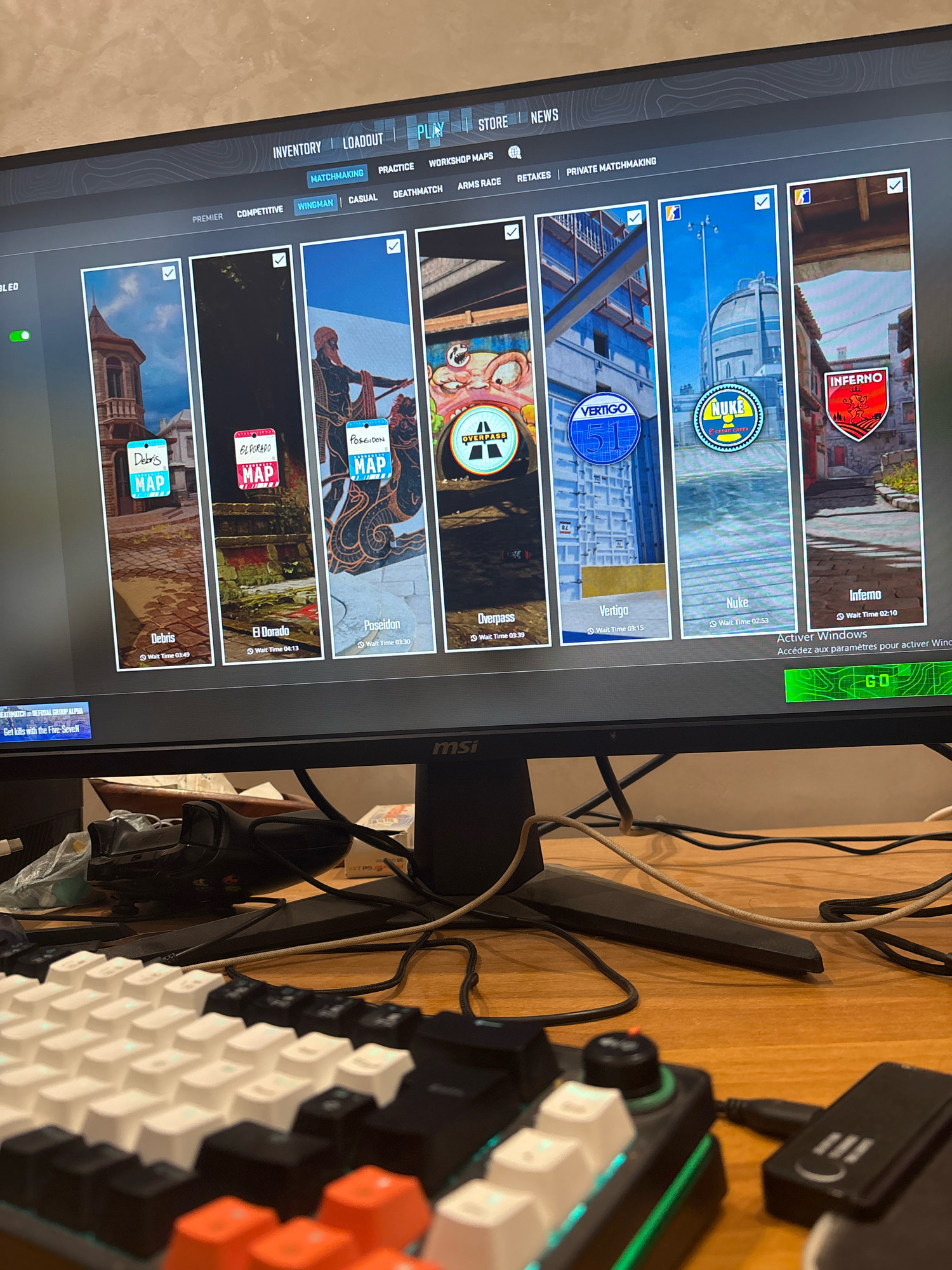 The image shows a computer monitor displaying a video game menu. The game appears to be a first-person shooter with a selection of maps and game modes. The user interface includes options for "Inventory," "Loadout," "Play," "Store," and "News." Below these are various game mode selections such as "Matchmaking," "Practice," "Workshop Maps," "Premier," "Competitive," "Wingman," "Casual," "Deathmatch," "Arms Race," "Retakes," and "Private Matchmaking." The central part of the screen shows a carousel of available maps with their names and estimated wait times: "Debris" (Wait Time 03:49), "El Dorado" (Wait Time 04:13), "Poseidon" (Wait Time 03:30), "Overpass" (Wait Time 03:39), "Vertigo" (Wait Time 03:15), "Nuke" (Wait Time 02:53), and "Inferno" (Wait Time 02:10). A green button labeled "GO" is visible at the bottom right of the screen, along with the text "Activer Windows" which is French for "Activate Windows." The monitor is situated on a wooden desk with a keyboard in the foreground. Cables and computer peripherals are also visible on the desk. The background is out of focus, but appears to be an indoor setting. There are no people visible in the image. The image does not contain any visual elements that indicate a location in the city of Marrakesh, Morocco.
