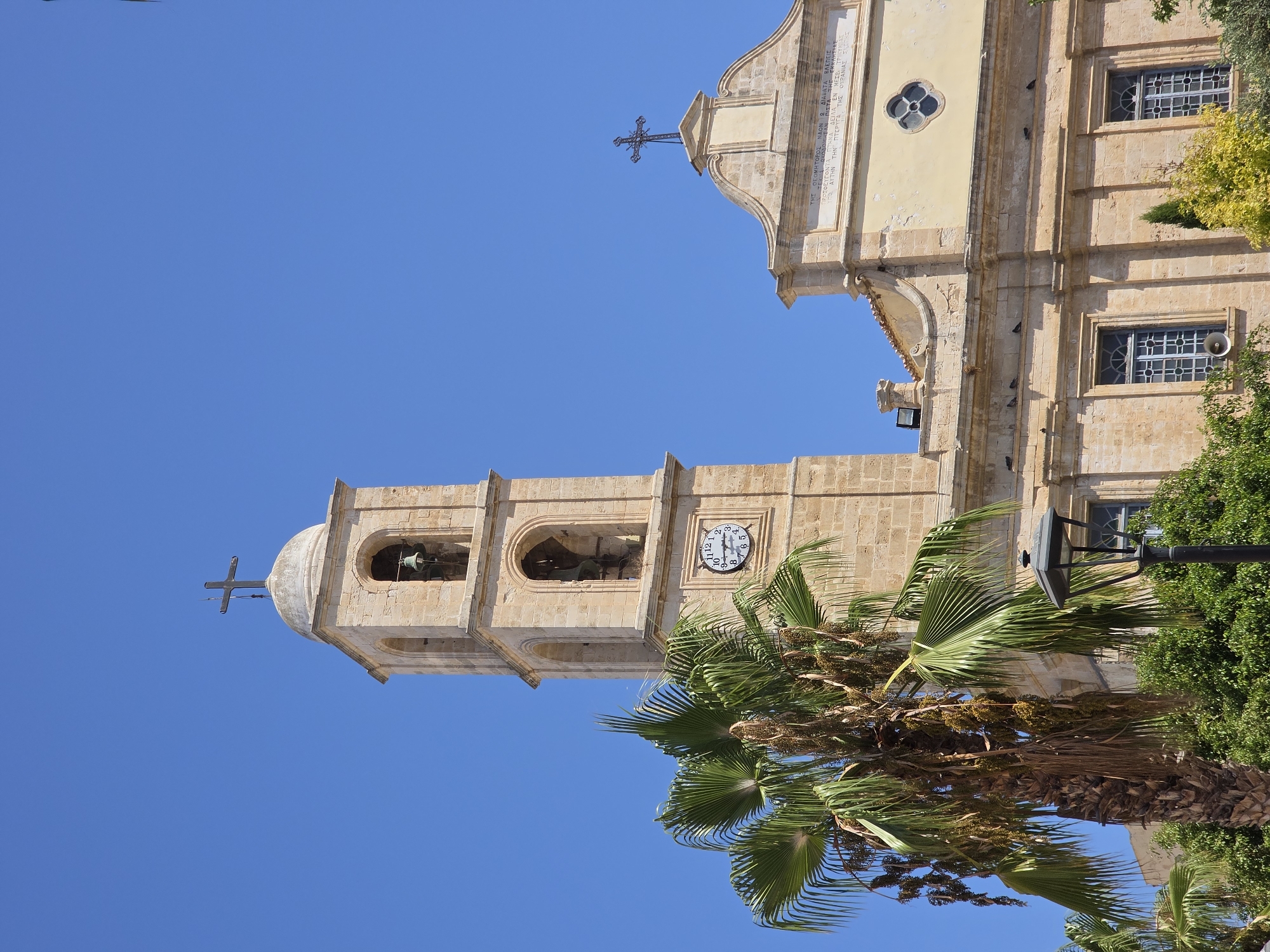 This image captures a view of the Church of Saint Titus in Chania, Greece. The church features a prominent bell tower made of stone, topped with a dome and a cross. The tower has arched openings revealing bells and a clock face showing approximately 10:08. Adjacent to the bell tower is the main body of the church, constructed from similar stone and painted a light yellow. This section has a decorative facade with a large inscription above the entrance, a cross, and a circular window. A large palm tree with drooping fronds stands in the foreground, obscuring part of the church's lower section. Other foliage and a black lamppost are also visible. The sky is a clear, bright blue.