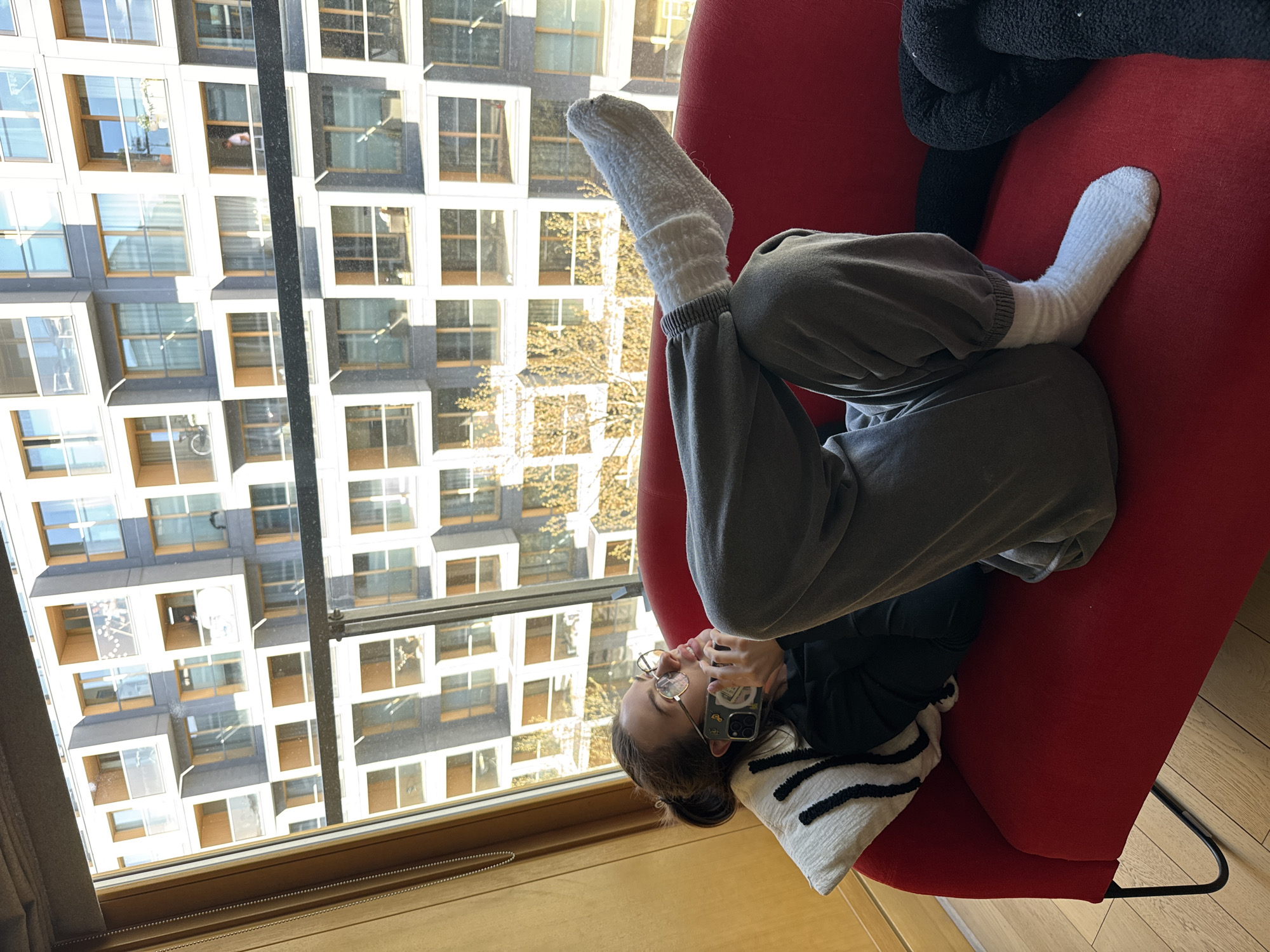 A young adult female is lying on a red sofa in a well-lit room, possibly an apartment. She is wearing gray sweatpants and white socks, and she is holding a smartphone up to her ear with her left hand. She has glasses on and is resting her head on a white pillow with black stripes. In the background, a large window shows a view of a building with many windows. Some windows contain potted plants and other items. Based on the architectural style and urban setting visible through the window, the location is most likely in the city of Warsaw, Poland. The apartment has wood floors. The angle suggests the picture was taken overhead, looking down.