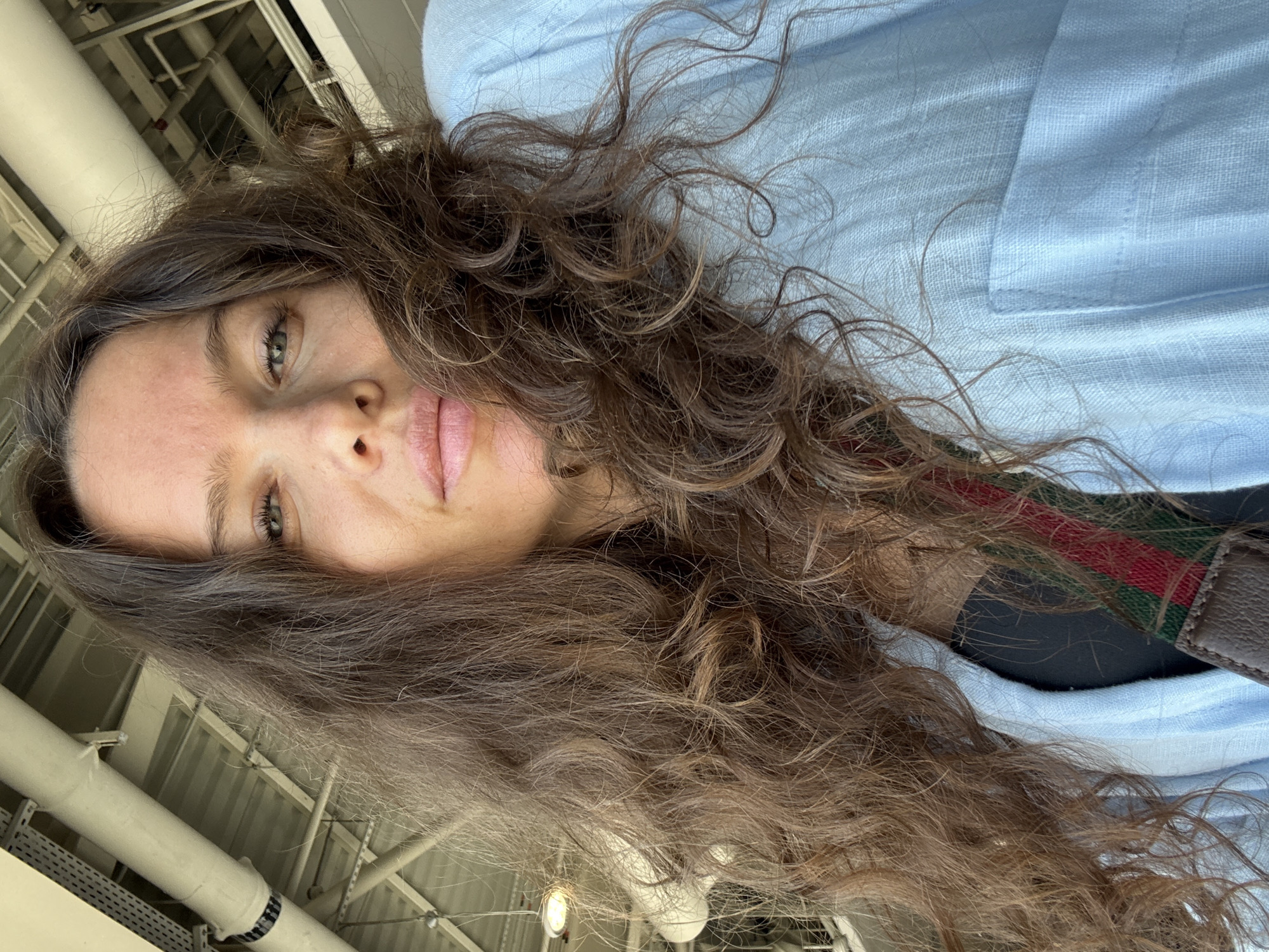 The image is a close-up, upward-facing selfie of a woman with long, wavy brown hair. She is wearing a light blue shirt and a black top underneath, with a strap that appears to be part of a bag featuring red and green stripes. Her skin is fair, and she has green eyes. The background shows an industrial ceiling with exposed pipes and lighting fixtures, characteristic of a large indoor venue or a covered outdoor area. The lighting suggests it is daytime. The overall environment appears to be a public space with utilitarian architecture. There is no specific indication of location within Dubai, United Arab Emirates.