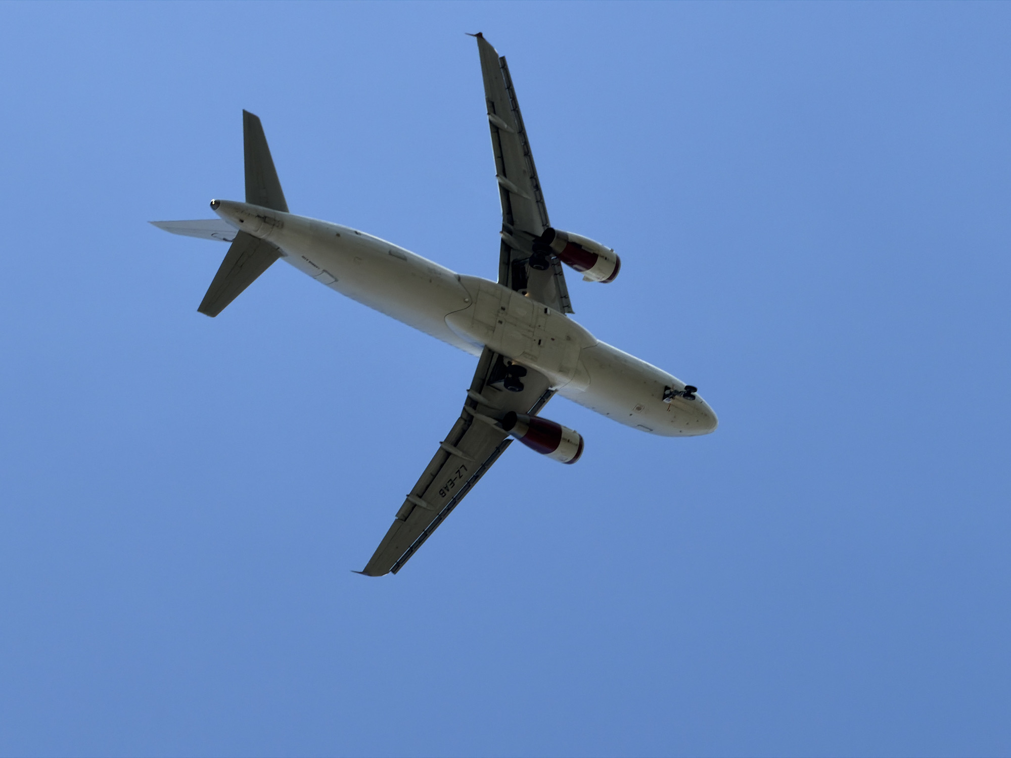 An overhead shot of a white commercial airplane in flight against a clear blue sky. The airplane's underside is visible, showing its wings with engines attached, the fuselage, and the tail. The registration mark "LZ-FAB" is visible on the wing. The landing gear is deployed. The photograph was taken in Burgas, Bulgaria.