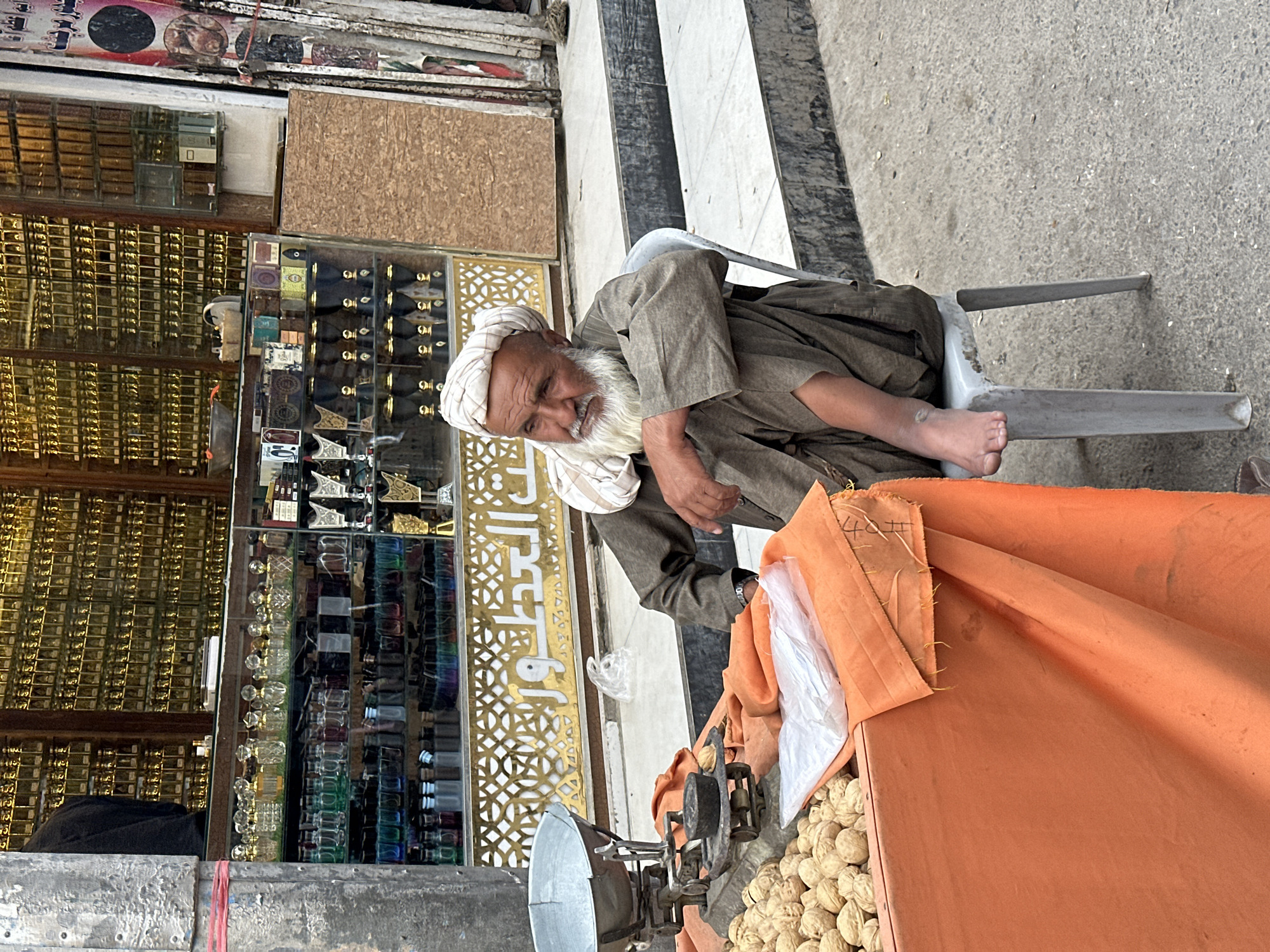 A man is sitting on a grey plastic chair on a street in Quetta City Tehsil, Pakistan. The man is elderly with white hair and a beard, wearing a tan turban and a long-sleeved robe. To his right is an orange draped table with walnuts on it and a vintage weighing scale. Behind the man is a store with glass display cases, filled with small glass bottles and containers. The store facade is decorated with a gold patterned design. The street has a black and white tiled section and a section of concrete.