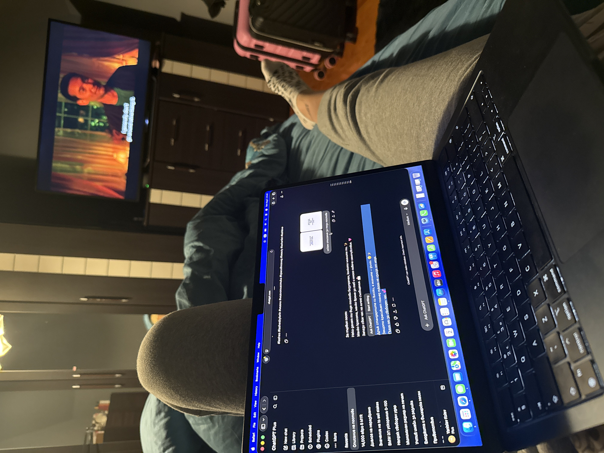 In a dimly lit bedroom, a person's legs are visible, covered in gray sweatpants, resting on a blue duvet. A MacBook laptop, displaying the ChatGPT interface, is positioned on the bed in front of the person. On the laptop screen, there are Bulgarian characters, indicating the language of the text. To the right of the laptop, a television screen shows a scene from a movie or TV show, featuring an Asian man. In the background, a wardrobe with mirrored doors is visible, along with two suitcases, one pink and one black. The overall ambiance suggests a relaxed evening at home, possibly in the city of Shumen, Bulgaria, given the Bulgarian text on the laptop.