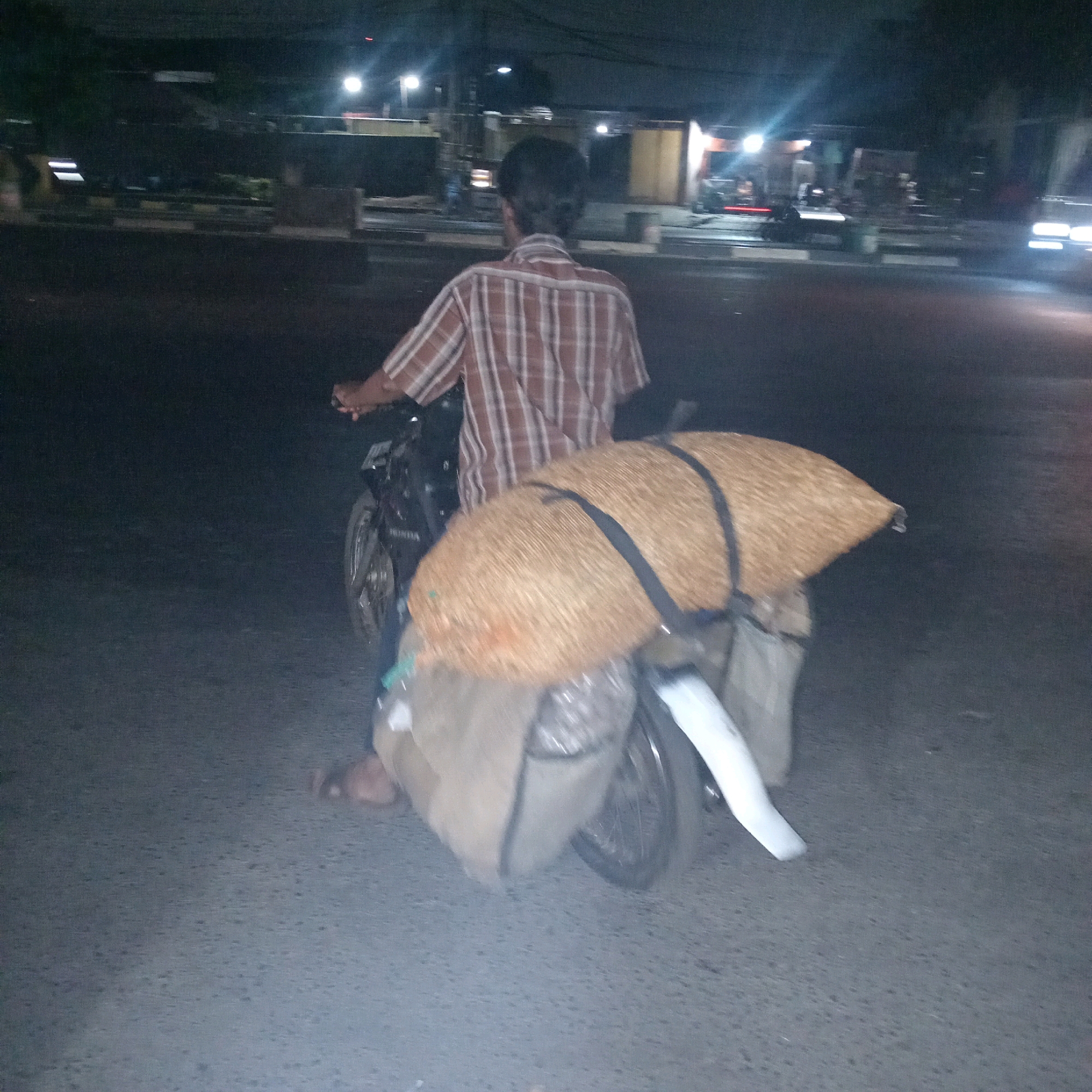 A man wearing a brown and white plaid shirt is riding a motorcycle at night in Cileungsi, Indonesia. The motorcycle is carrying a large amount of cargo, including a large straw-colored sack secured with black straps and two large, light-colored bags underneath. The man is seen from behind, and his hands are on the handlebars. The environment appears to be a street at night, with streetlights and buildings visible in the background, suggesting an urban or semi-urban setting. The road surface is asphalt.
