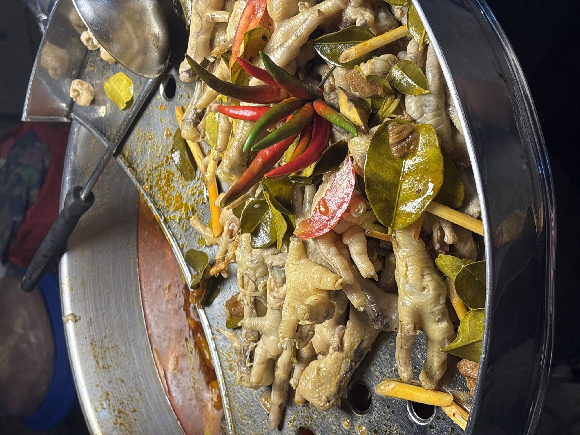 A close-up, top-down shot reveals a large, round, metallic cooking pot or steamer filled with cooked chicken feet, seasoned with various herbs and spices. The primary ingredients visible are numerous pale-skinned chicken feet, bright red and green chili peppers arranged centrally, and glossy green kaffir lime leaves scattered throughout. There are also slender yellow or orange stalks, possibly lemongrass or turmeric, and pieces of what appear to be red tomato or bell pepper.
The pot itself is made of metal, showing some wear and discoloration from use, with a reddish-brown liquid visible at the bottom on the left side, suggesting a simmering sauce or broth. A metal ladle is resting on the rim of the pot on the left, indicating that the food is either being prepared for serving or kept warm for consumption. The lighting is dim and artificial, suggesting a nighttime setting or an indoor environment, typical of a street food stall or a casual eatery. The dish and ingredients strongly suggest Southeast Asian cuisine, aligning with the location context of Choeng Thale, Thailand. No people or visible text are present in the image.